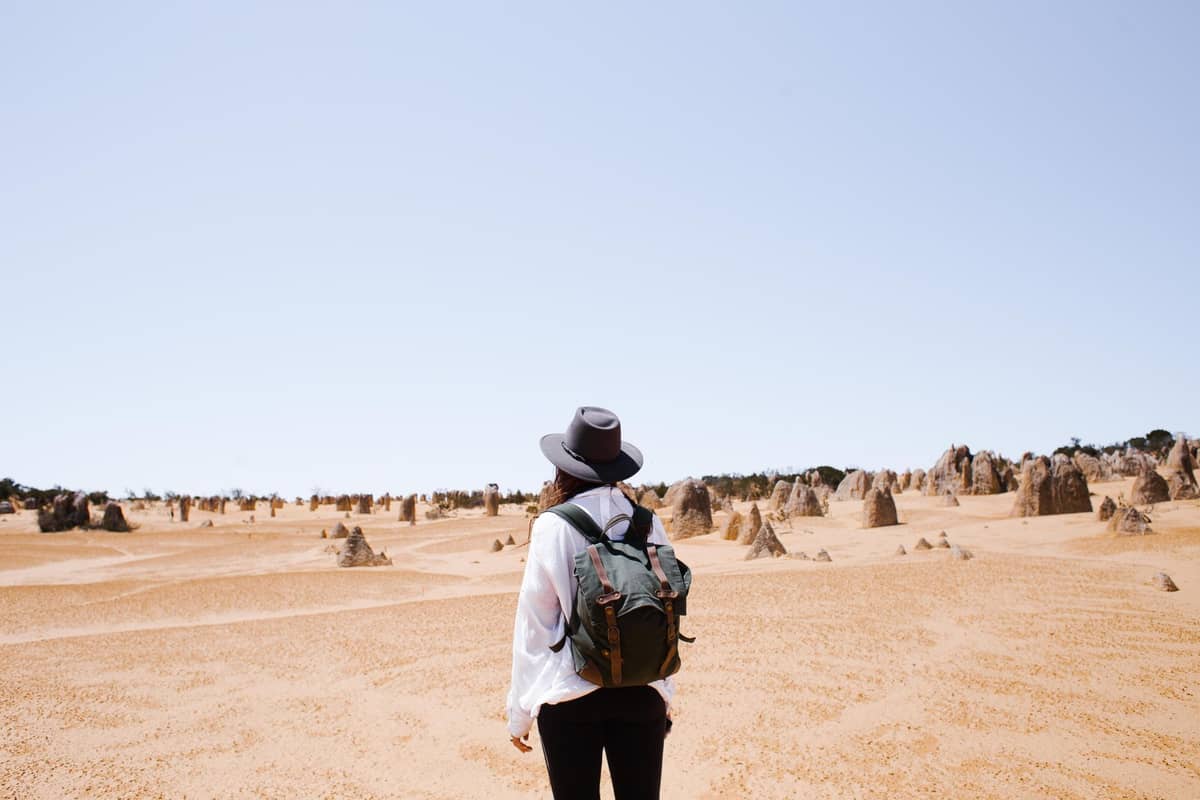 Person with backpack and hat stands looking out at a yellow sandy desert dotted with limestone pillars under a clear blue sky.