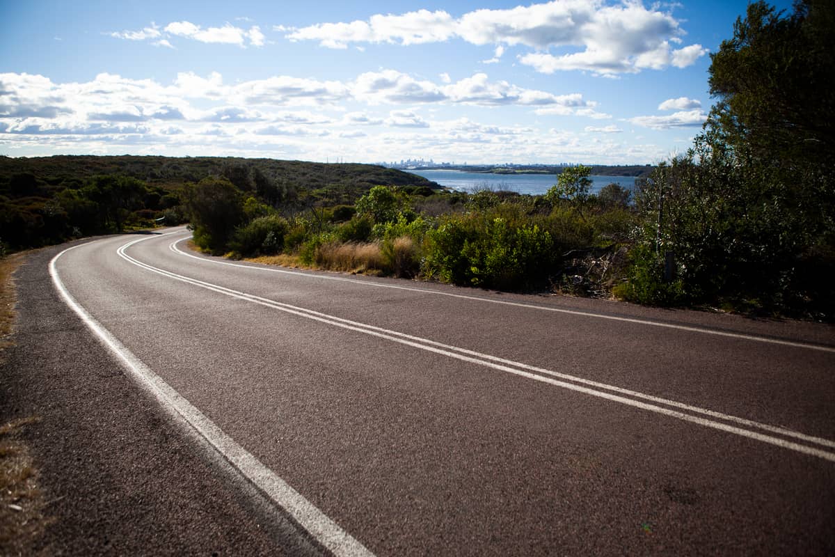 Sealed two-lane road curving through Australian bushland with distant water and blue sky, typical of scenic drives near Perth