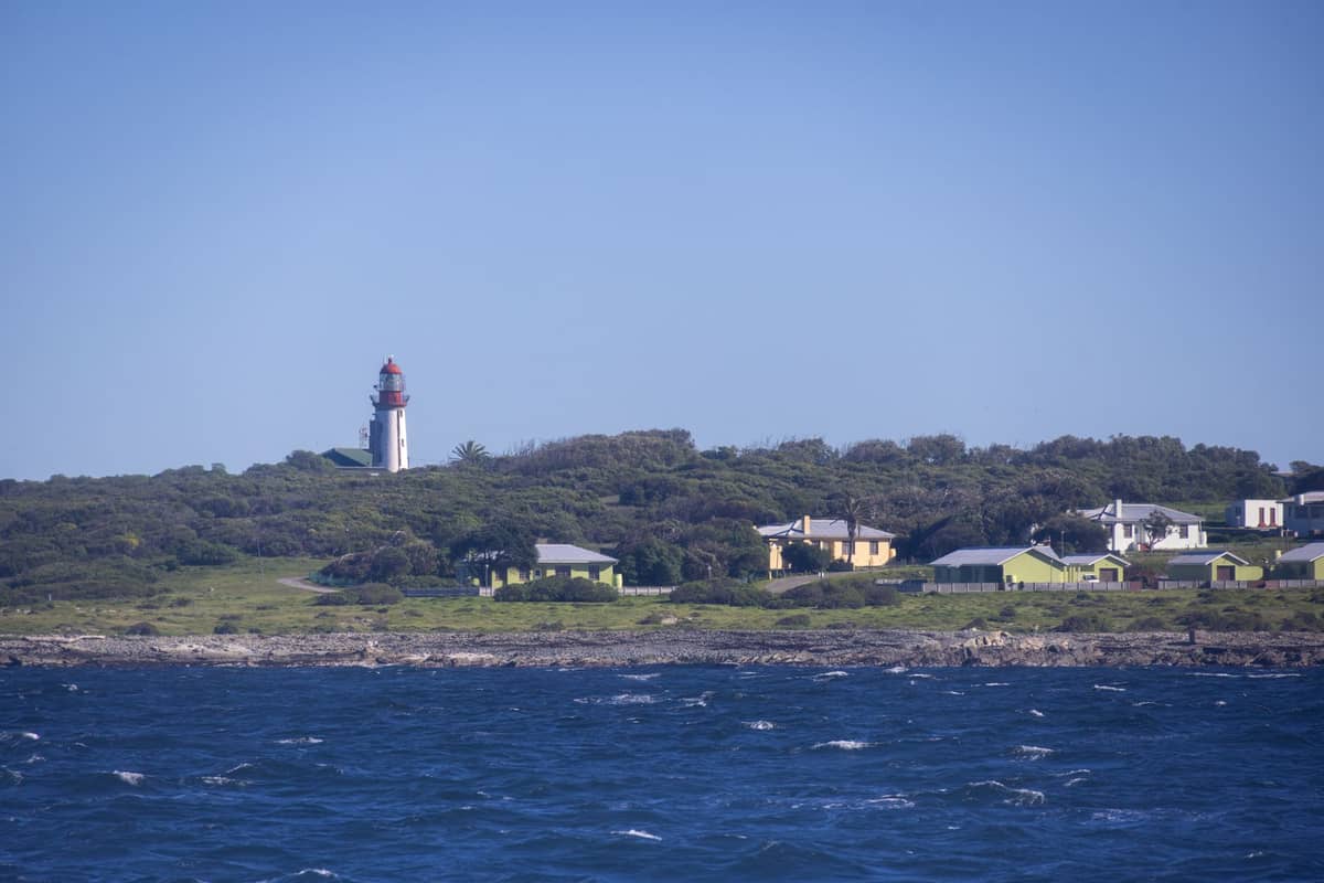 Blick auf die Küste mit einem Leuchtturm und kleinen Hütten auf einem flachen Hügel, vom Wasser aus unter einem klaren blauen Himmel gesehen.