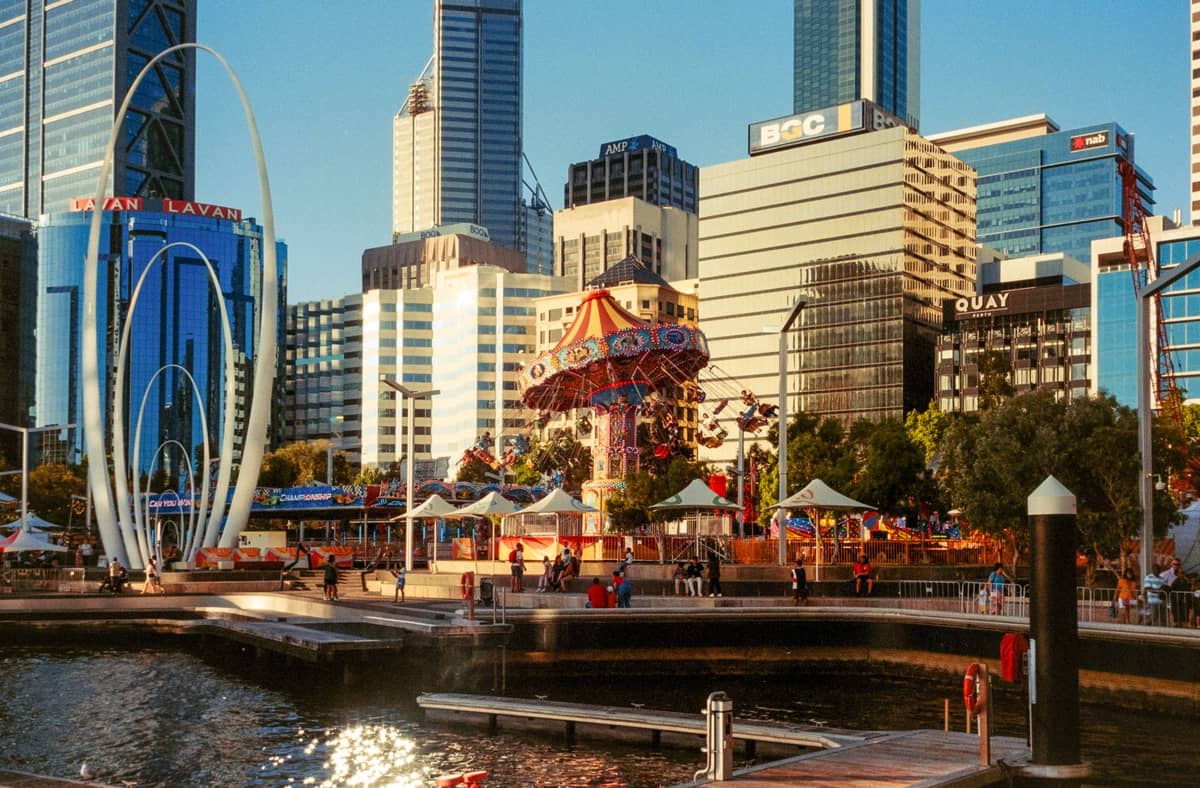 Elizabeth Quay in Perth in der Abenddämmerung mit Wolkenkratzern, Wasser und einem belebten öffentlichen Bereich mit Karussell und markanter Kunst im öffentlichen Raum.