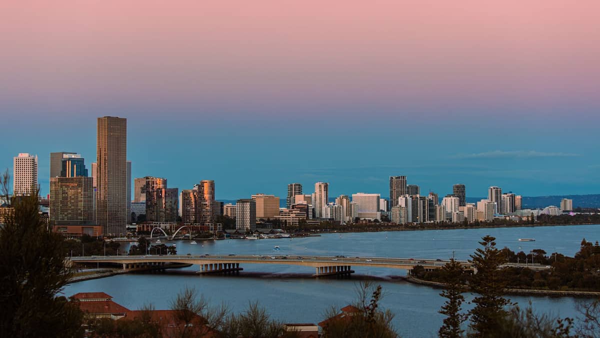 Weitwinkelansicht der Skyline des Perth CBD bei Sonnenuntergang über dem Swan River, mit Brücken, Stadtgebäuden und spiegelndem Wasser im Vordergrund.