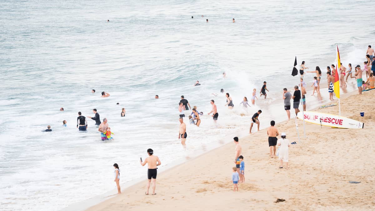 Belebter, bewachter Strand in Perth mit vielen Familien und Kindern, die im flachen Wasser in der Nähe eines Surf-Rettungsbretts und Sicherheitsfahnen spielen.