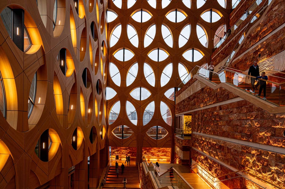 Innenansicht eines beeindruckenden zeitgenössischen Museumsraums mit geometrischen Fenstern, warmem Licht, Steinwänden und Familien, die die Ausstellungen erkunden.