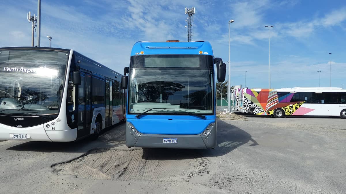Eine Reihe von Shuttlebussen mit der Aufschrift Perth Airport und bunten Grafiken, geparkt unter blauem Himmel auf einem Transportgelände.
