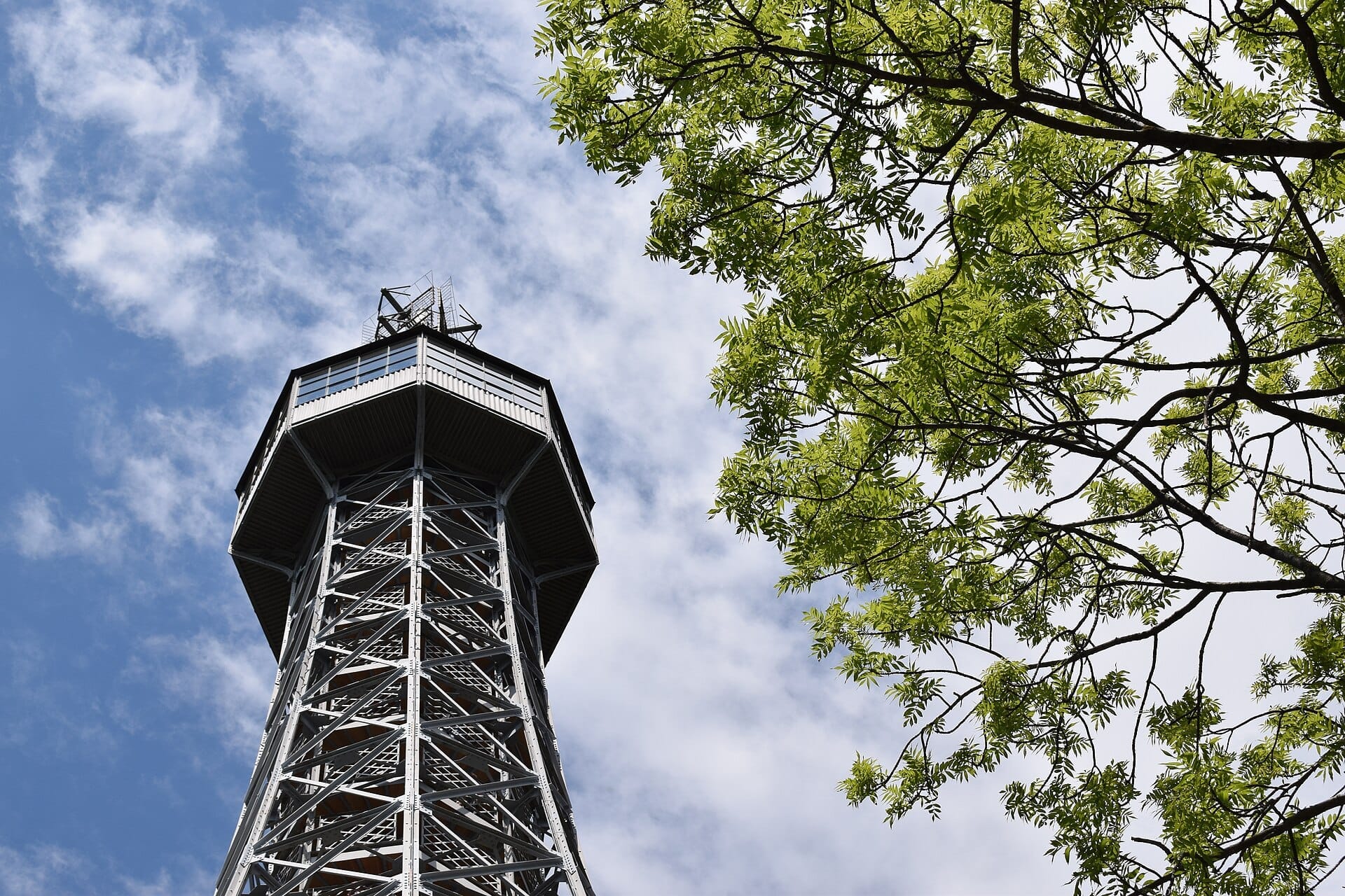 La Torre Belvedere di Petřín si erge contro un cielo blu parzialmente nuvoloso, incorniciata da fresche rami verdi nella luce primaverile.