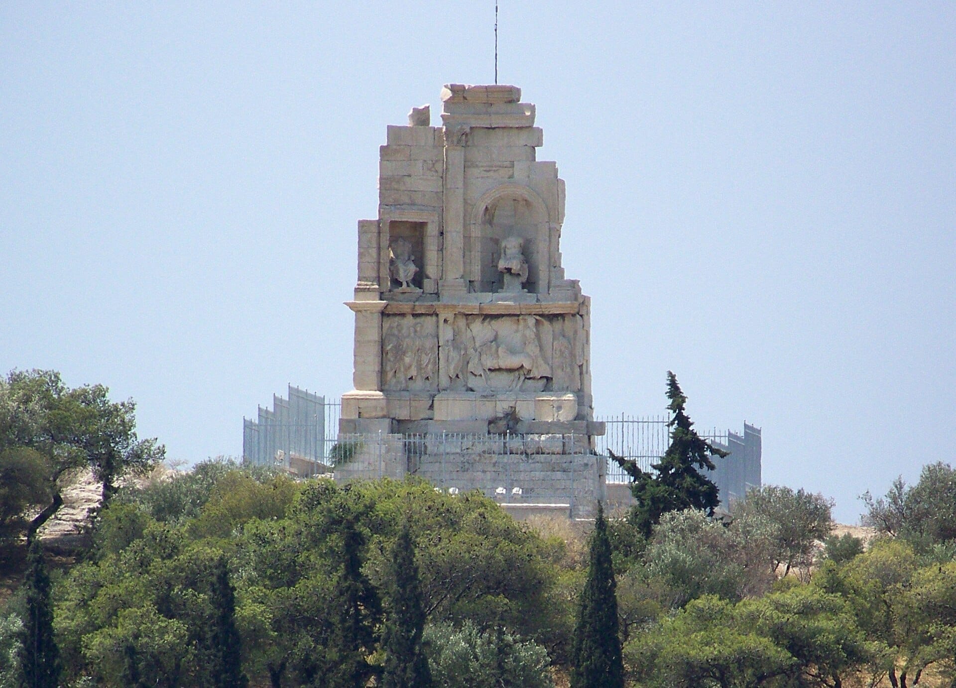 Il Monumento di Filopappo si eleva sopra pendii ricoperti di alberi sotto un cielo sereno, con i suoi antichi dettagli in marmo visibili da lontano sulla cima della collina.