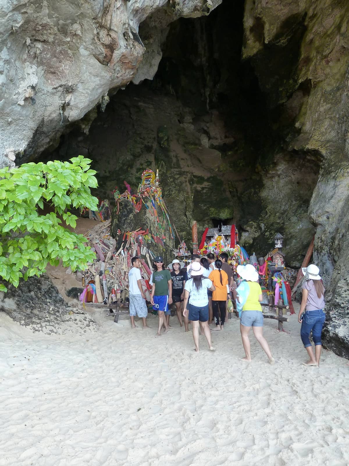 Besucher stehen auf sandigem Boden vor der Phra Nang Cave, umgeben von Kalksteinfelsen und bunten Holzopfergaben am Eingang des Prinzessinnen-Schreins.