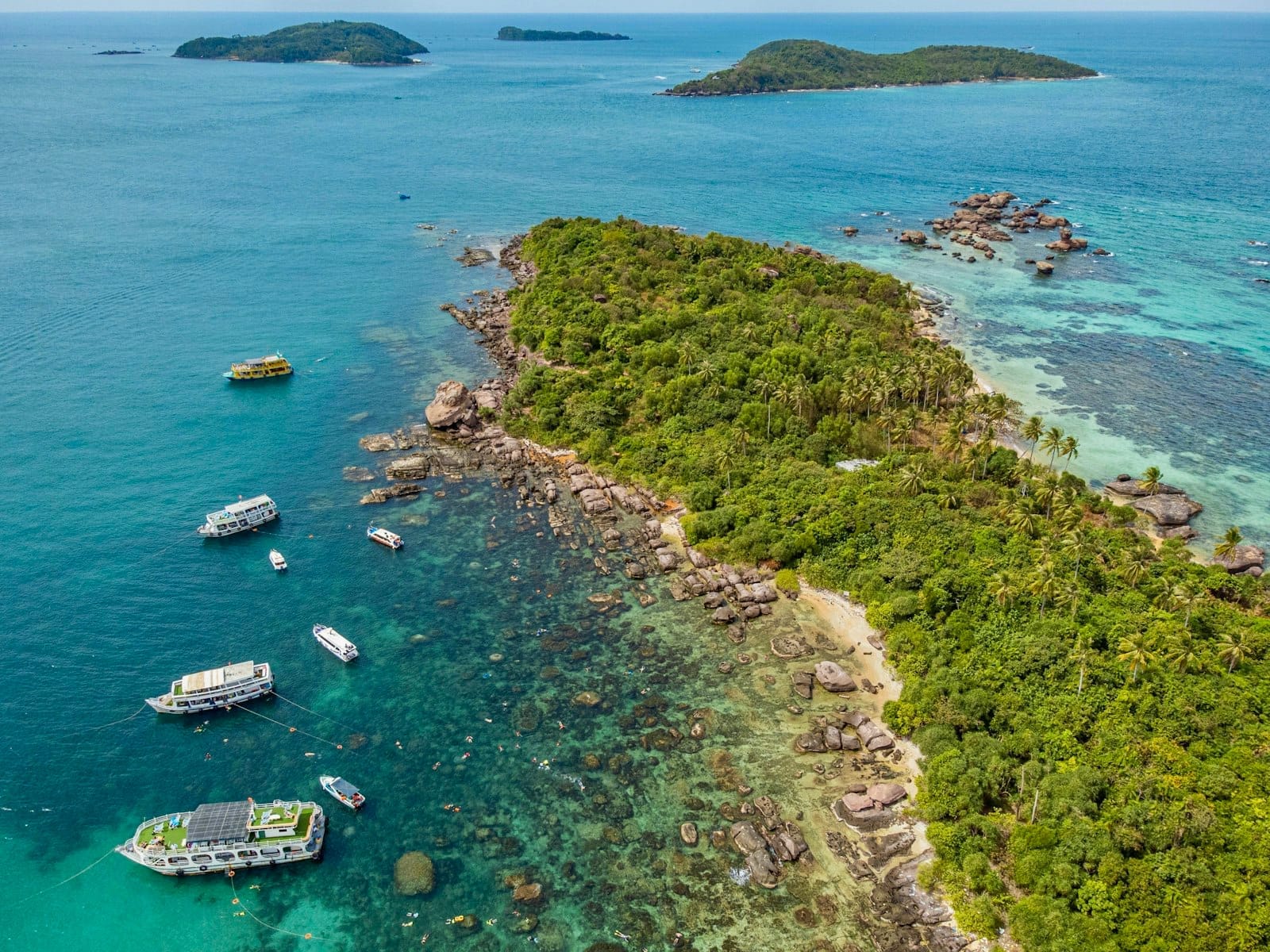 Luftaufnahme einer üppig grünen Insel mit vor der Küste ankernden Booten in klarem türkisfarbenem Wasser, typisch für Phu Quoc bei ruhigem Trockenzeit-Wetter.