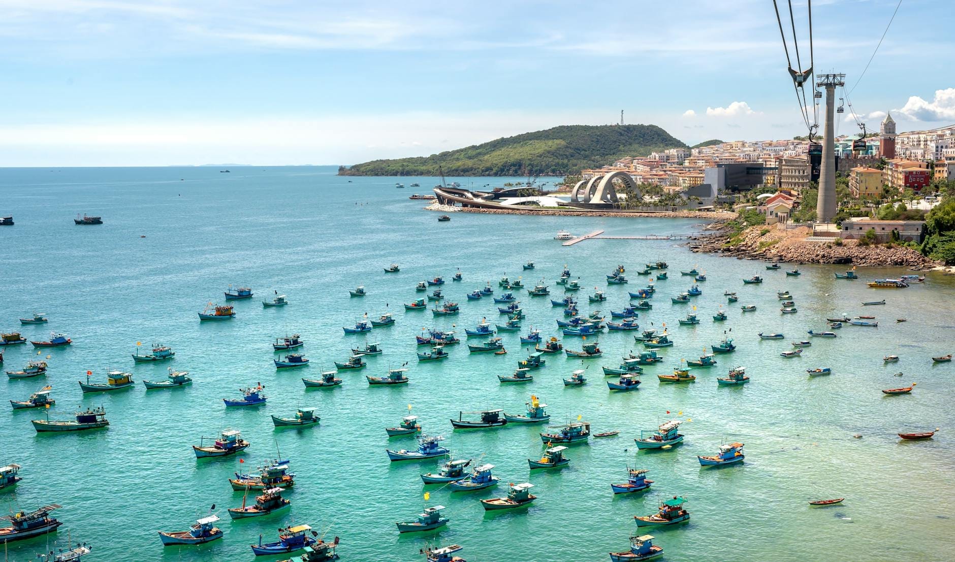 Bunte Fischerboote ankern im türkisfarbenen Wasser nahe An Thoi, im Hintergrund sind die Gondeltürme der Seilbahn und die Küstengebäude von Phu Quoc zu sehen.
