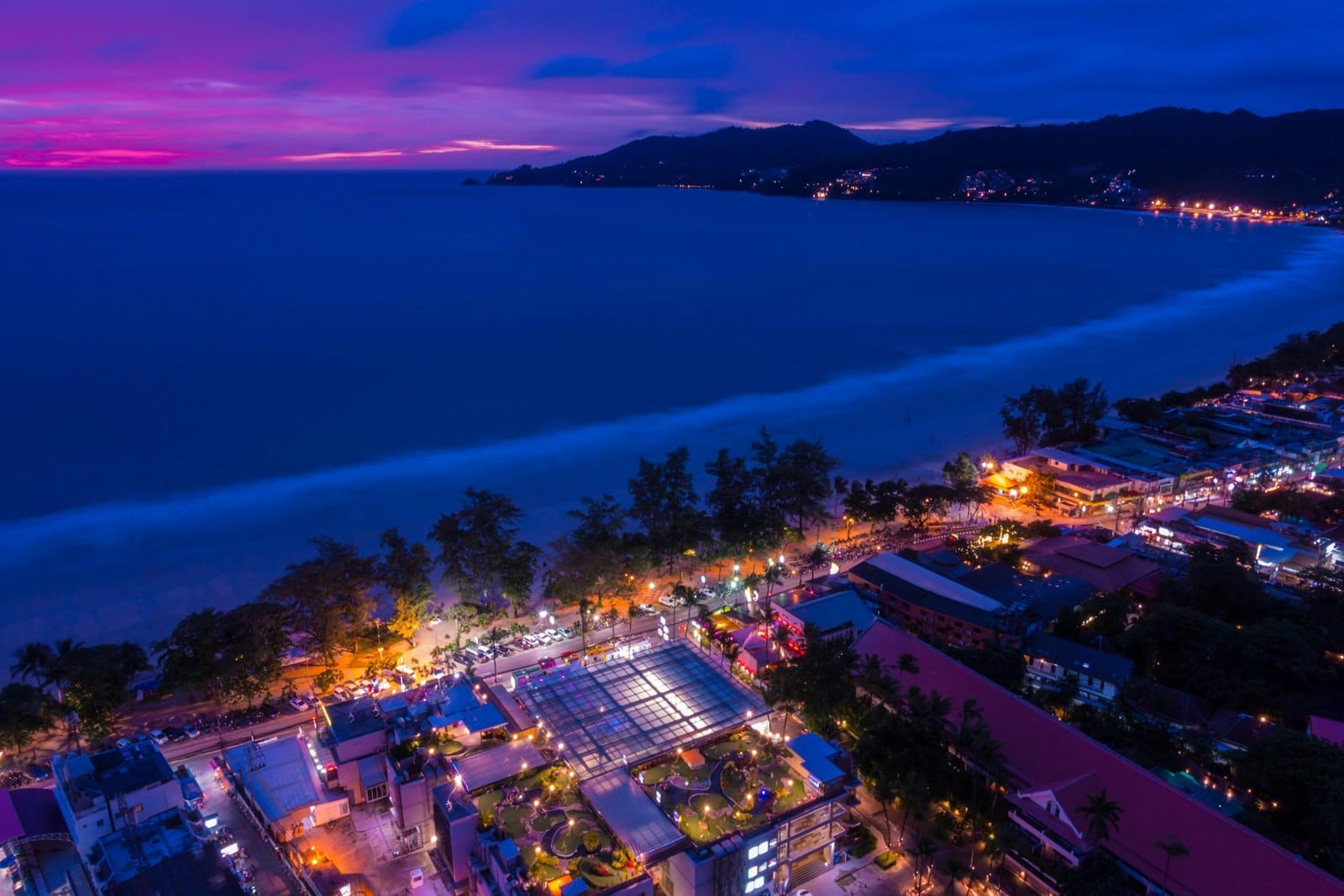 Aerial view of Patong Beach at dusk with lights from hotels and the city, and mountains in the distance.