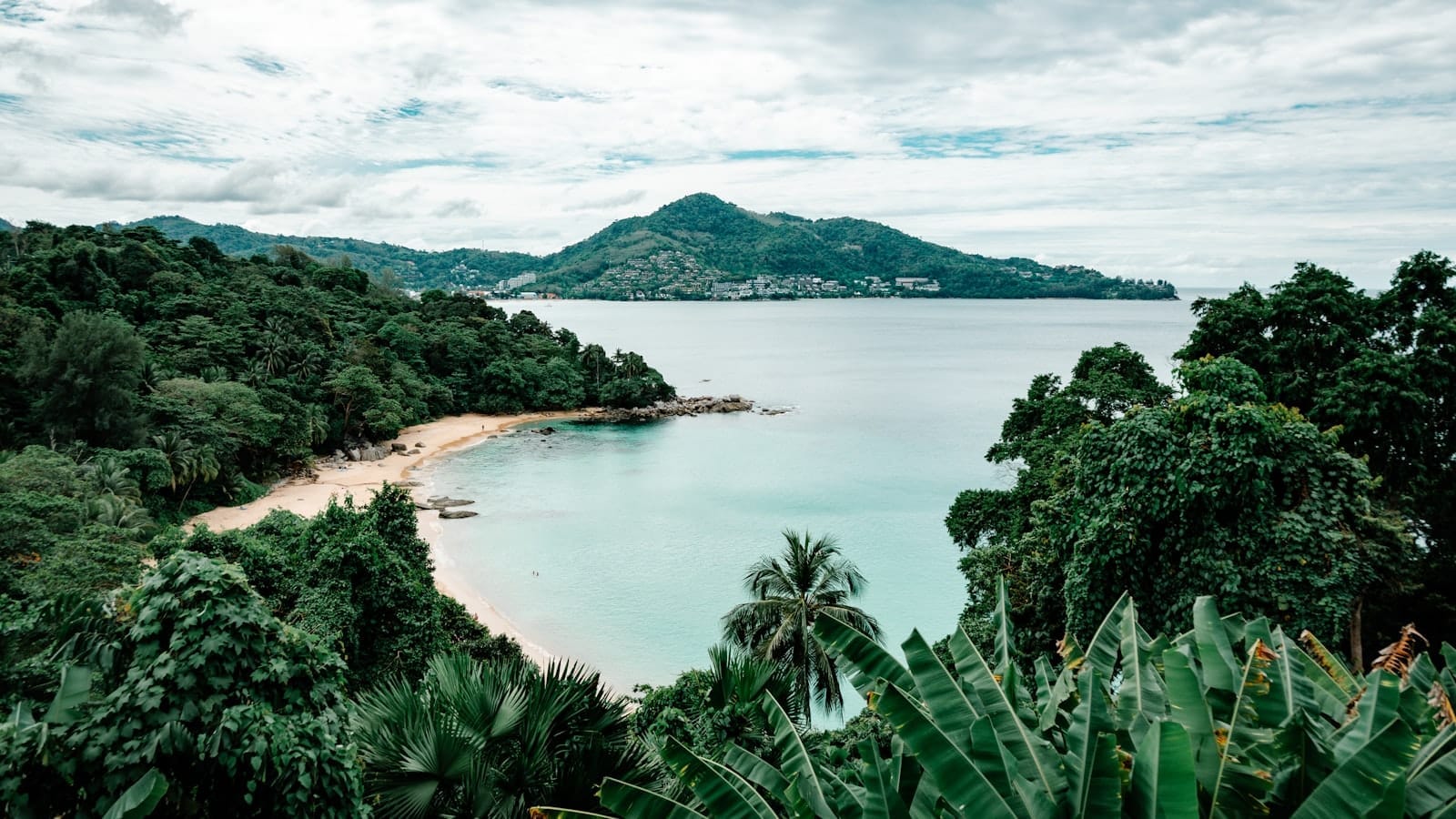 A panoramic view of a secluded Phuket beach surrounded by lush green hills, with turquoise sea and mountainous backdrop under a partly cloudy sky.