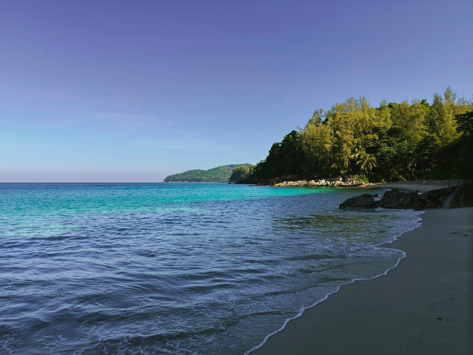 Wide, empty Phuket beach with turquoise water, forested headland, and no people, suggesting space and solitude.