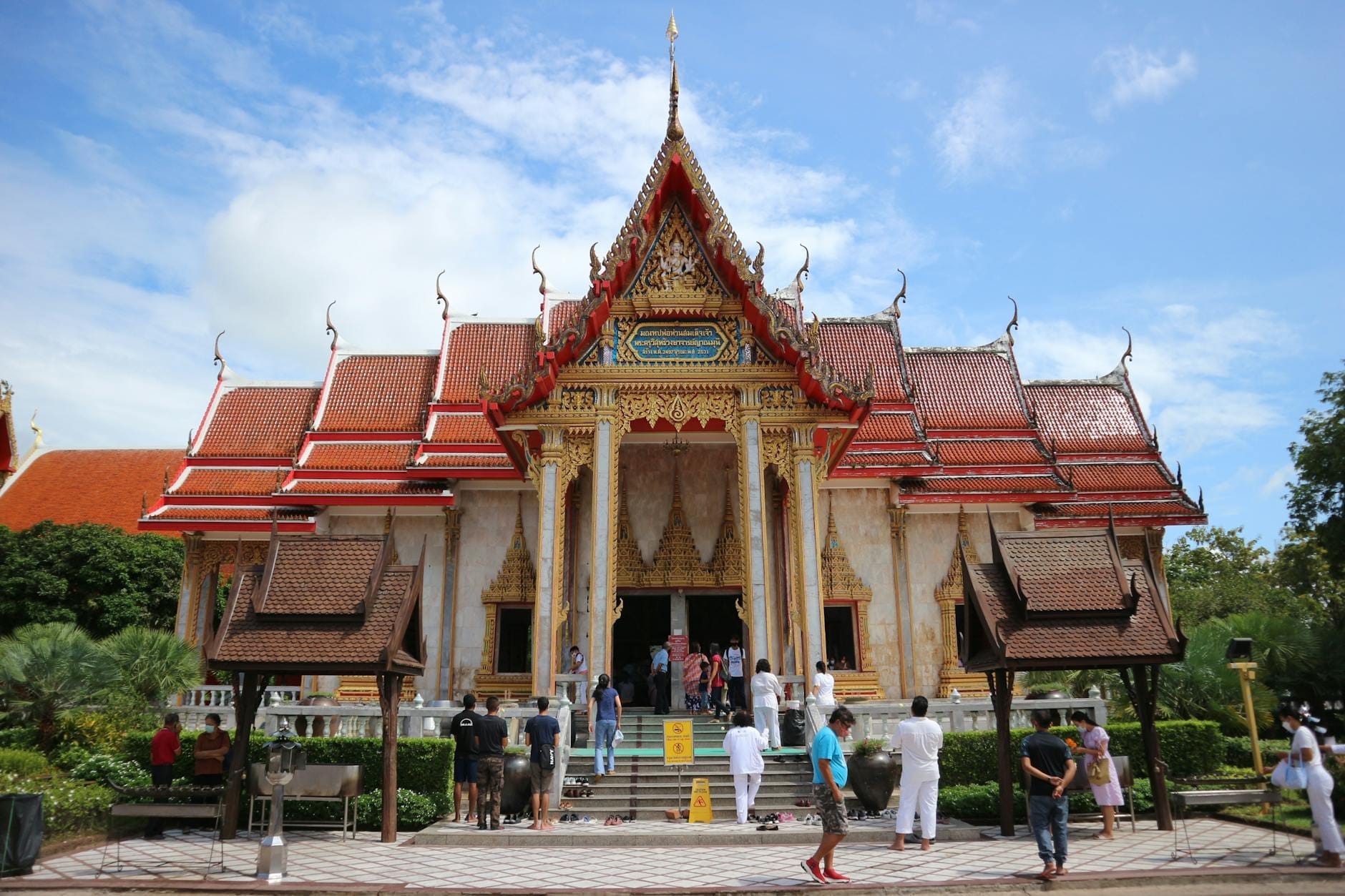 People visiting a large ornate Buddhist temple with red roof and gold accents, surrounded by greenery under a blue sky in Phuket.