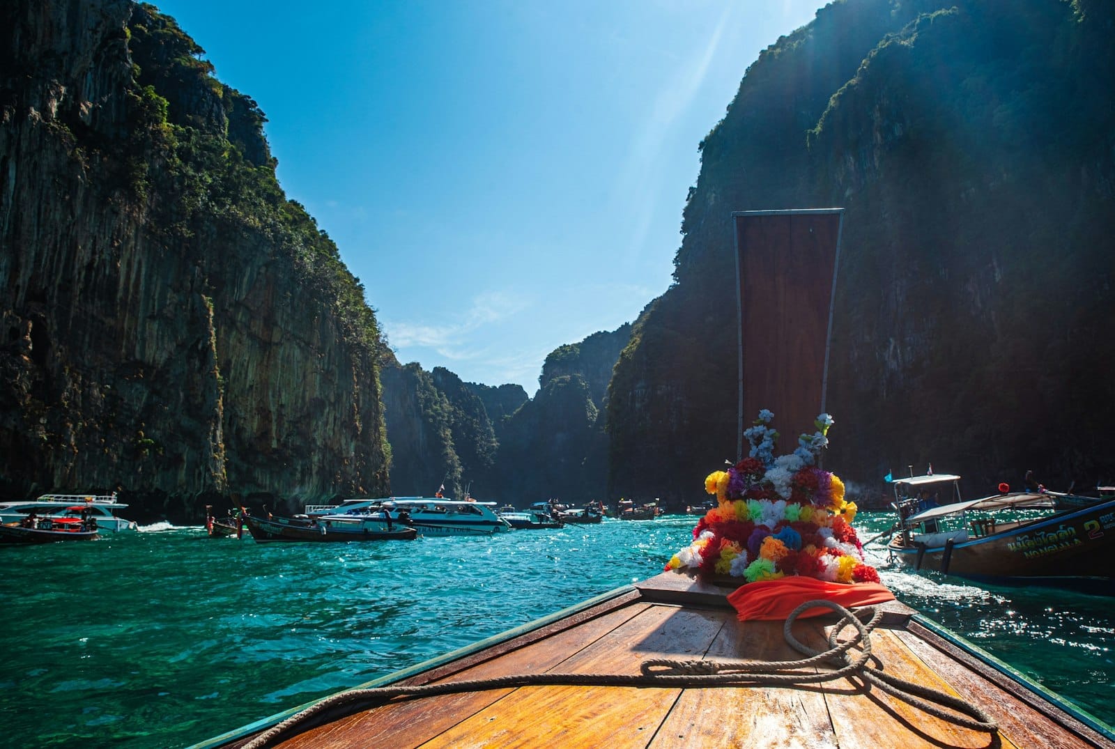 View from a traditional longtail boat approaching turquoise water and dramatic limestone cliffs, surrounded by other boats in a classic Phuket day trip scene.