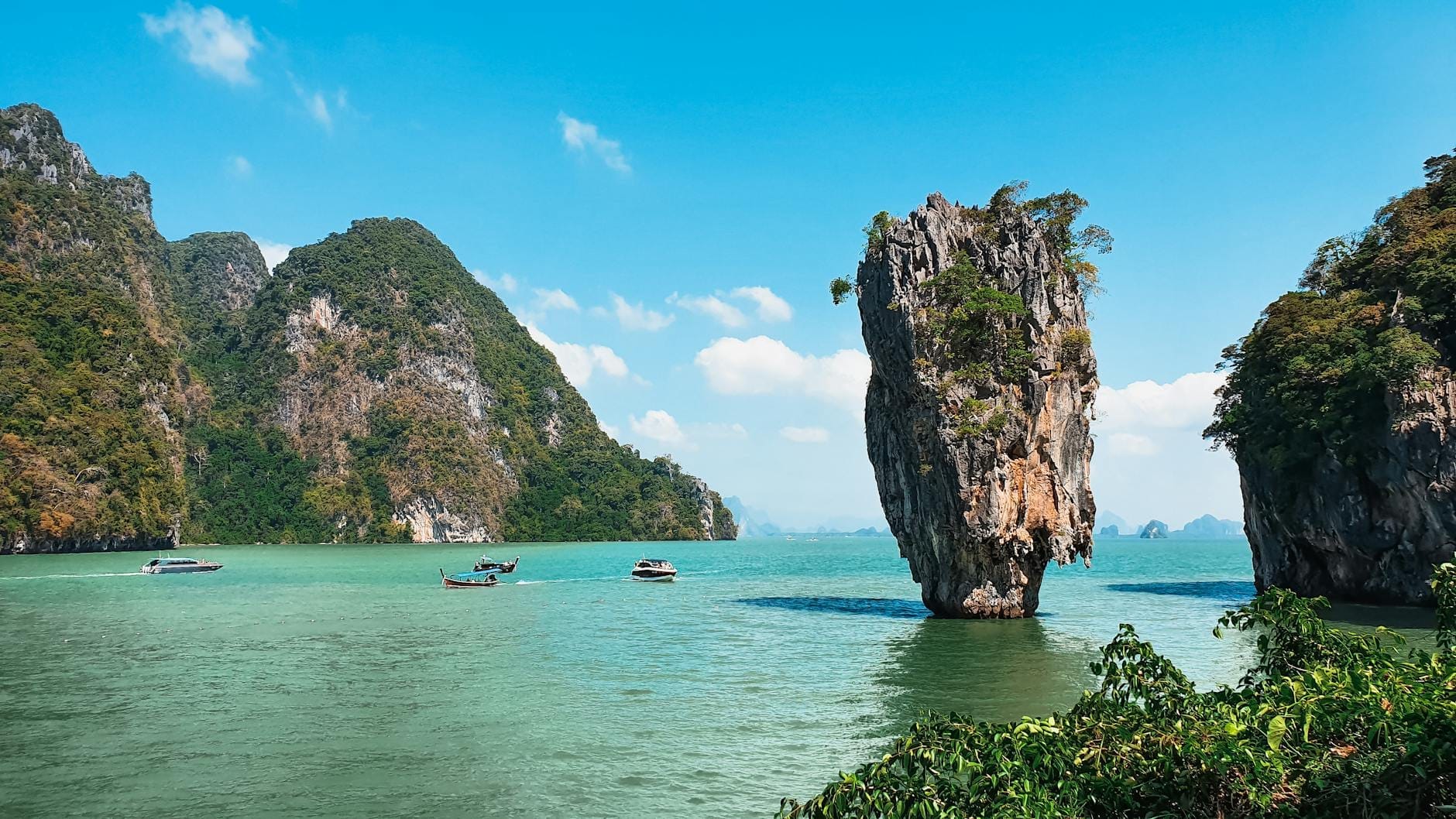 A bright view of James Bond Island with its iconic limestone rock in the sea, green hills, and clear blue sky, seen from the shore with boats nearby.