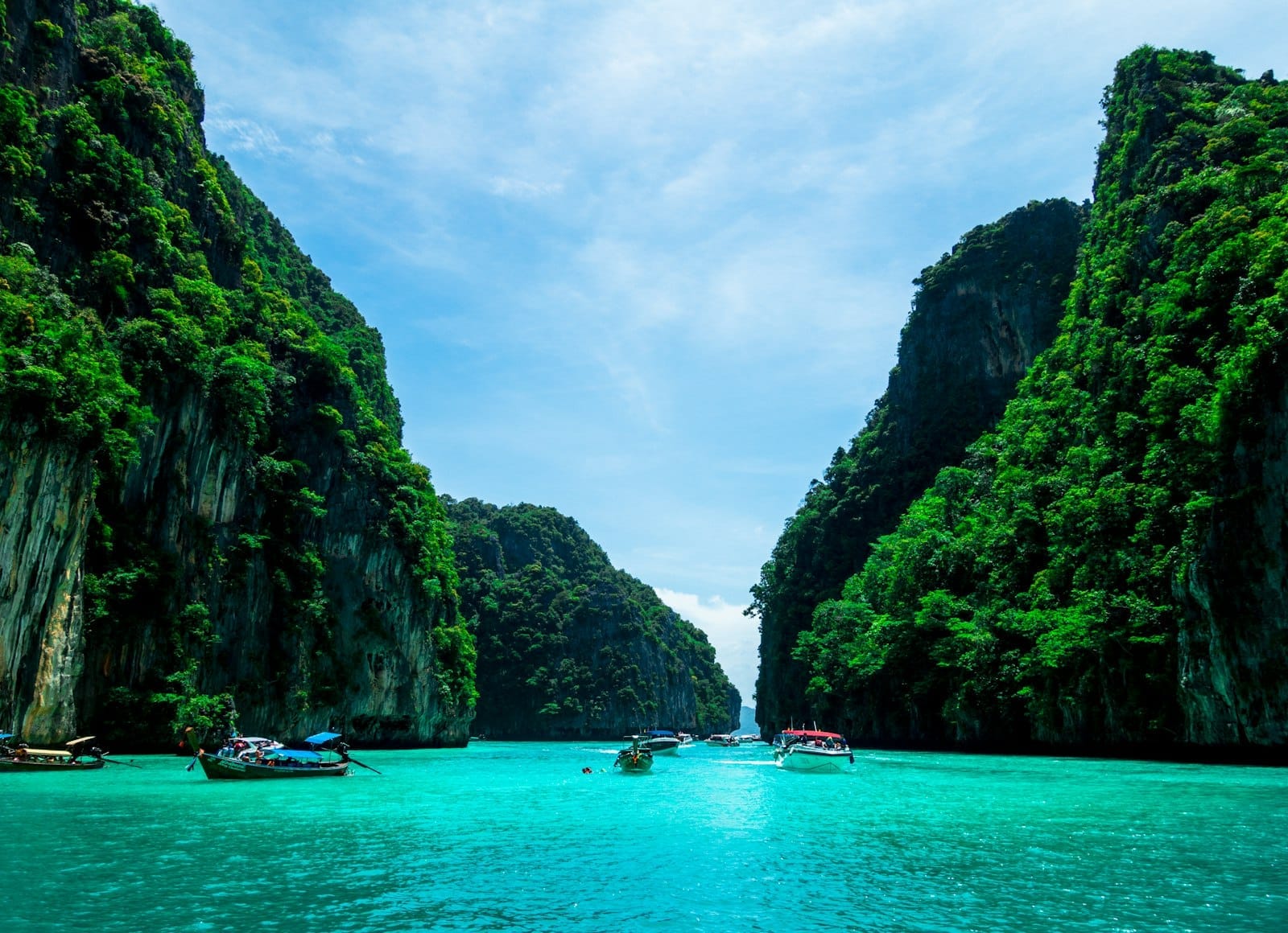 Turquoise waters surrounded by dramatic green cliffs with traditional longtail boats and speedboats, capturing the iconic tropical scenery of Phuket, Thailand on a sunny day.