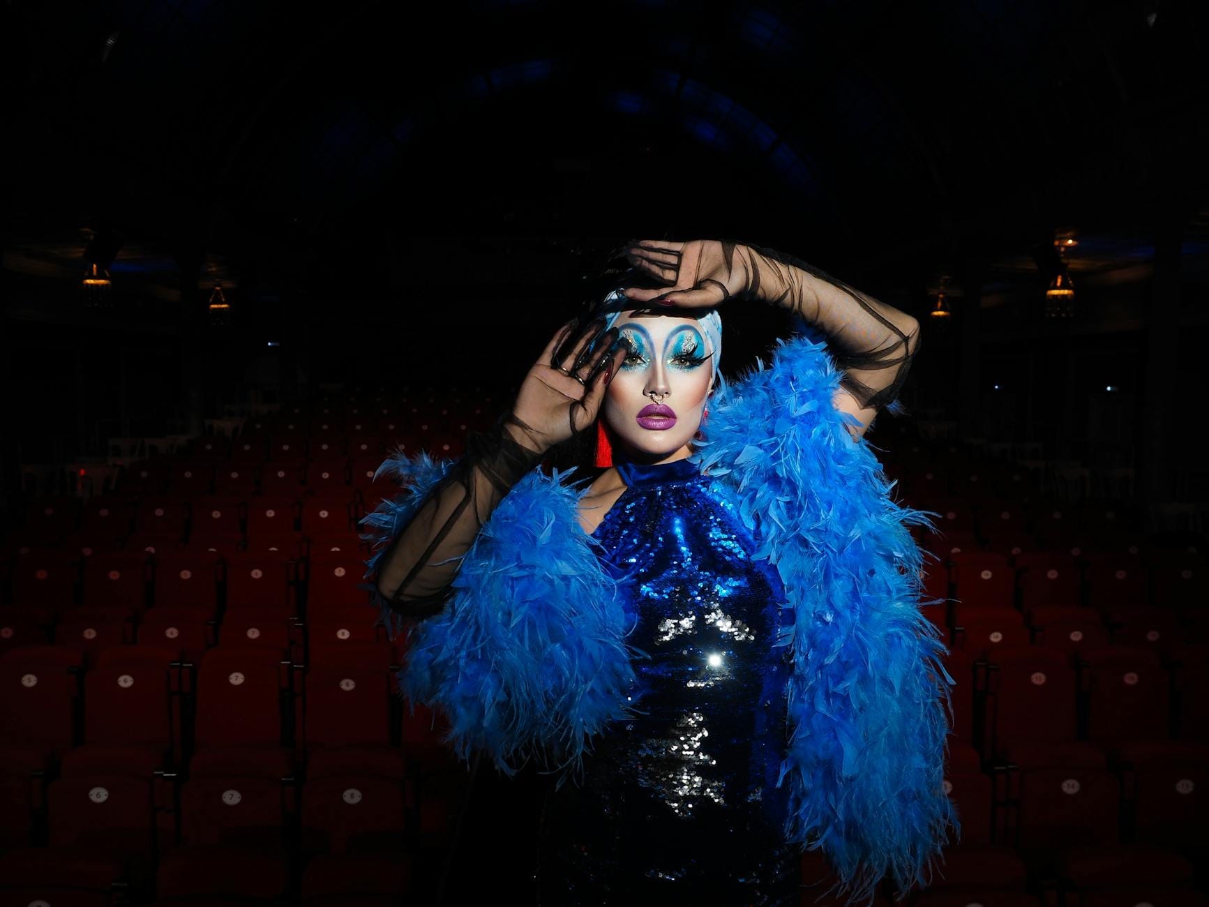 A flamboyantly dressed cabaret performer in a blue feathered costume and dramatic stage makeup poses in an empty theater.