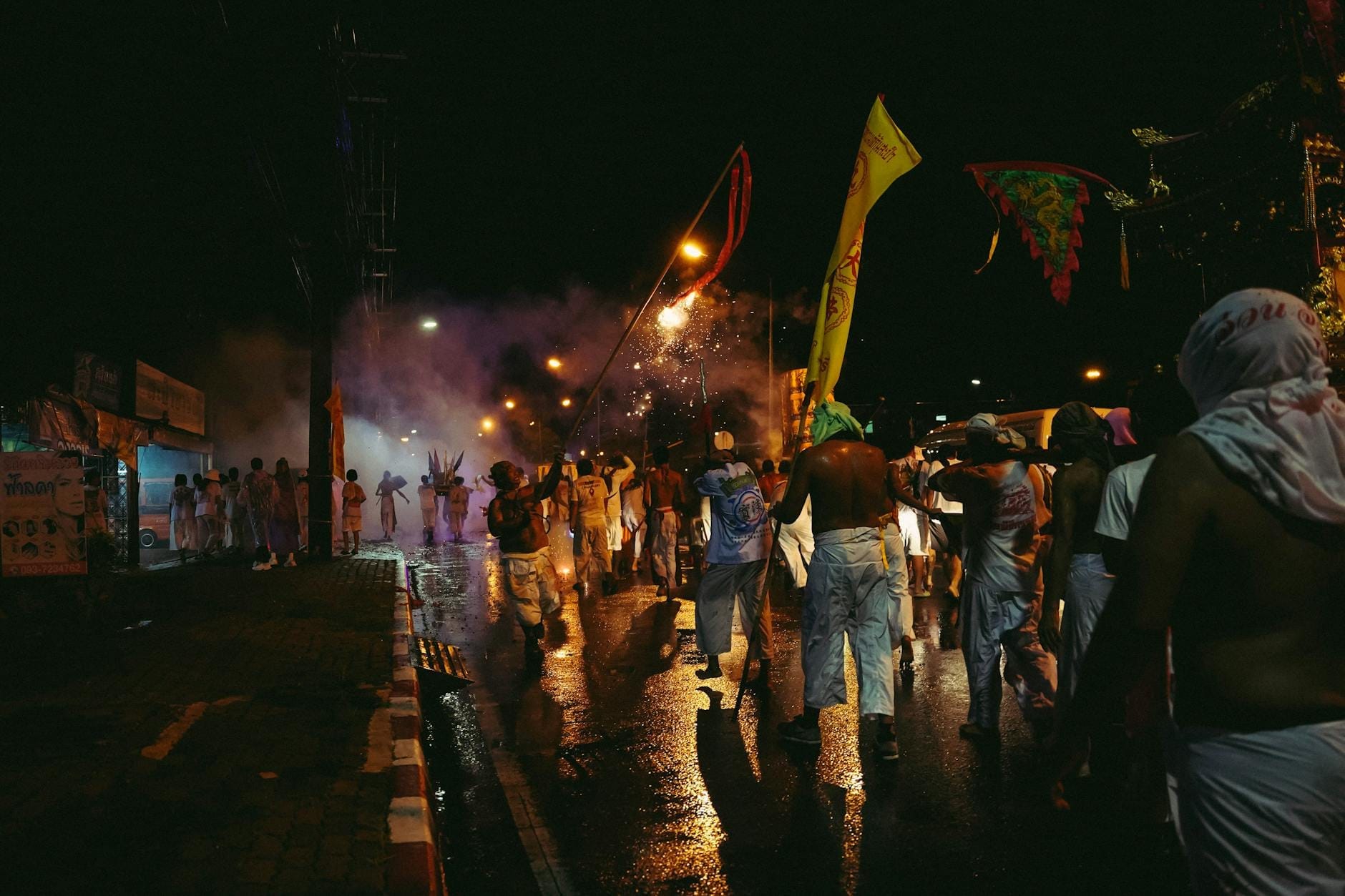 Nighttime street scene in Phuket with crowds and festive flags, people celebrating during a busy event under streetlights and smoky sky.