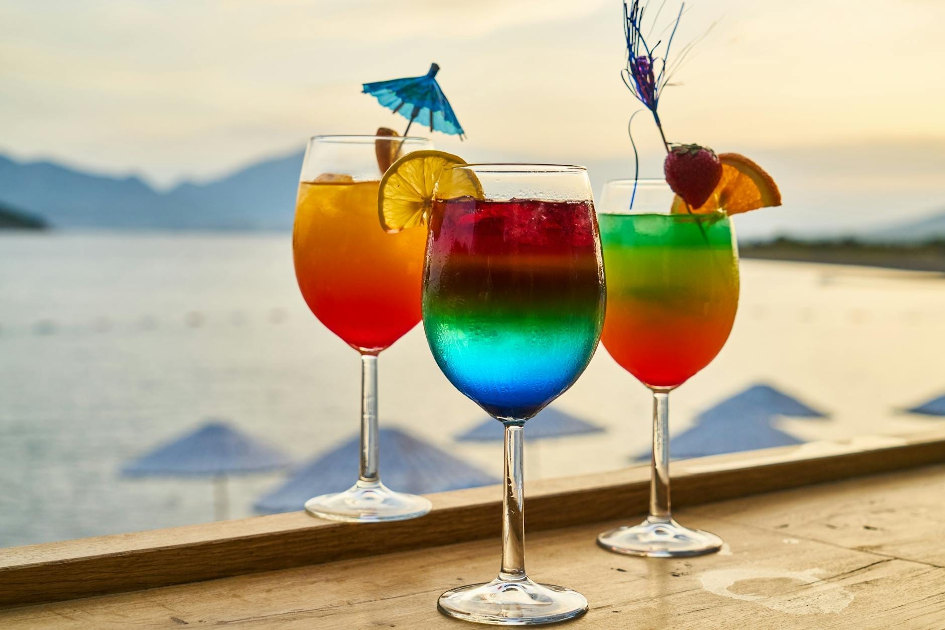 Three colorful cocktails with fruit garnishes on a wooden bar overlooking a beach and ocean at sunset with umbrellas in the background.