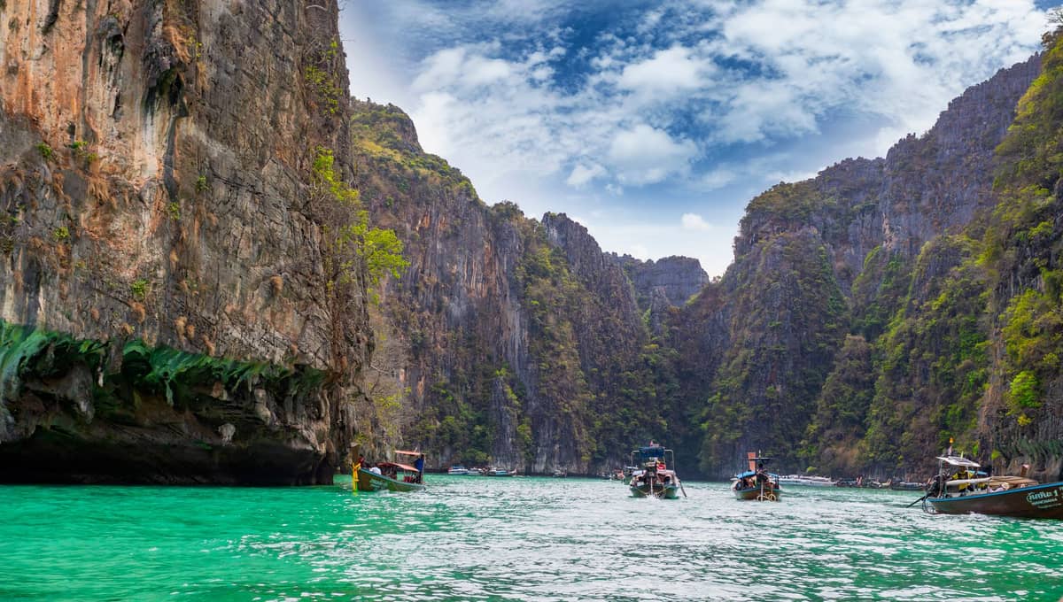 Blick vom Wasser auf die dramatischen Kalksteinfelsen in Krabi, Thailand, mit mehreren Longtail-Booten und Schnellbooten auf leuchtend türkisfarbenem Wasser.