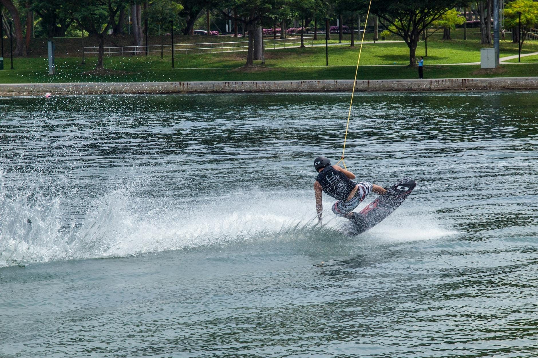Wakeboarder gliding across the lake at Phuket Wake Park, surrounded by green trees and grassy park under bright daylight.