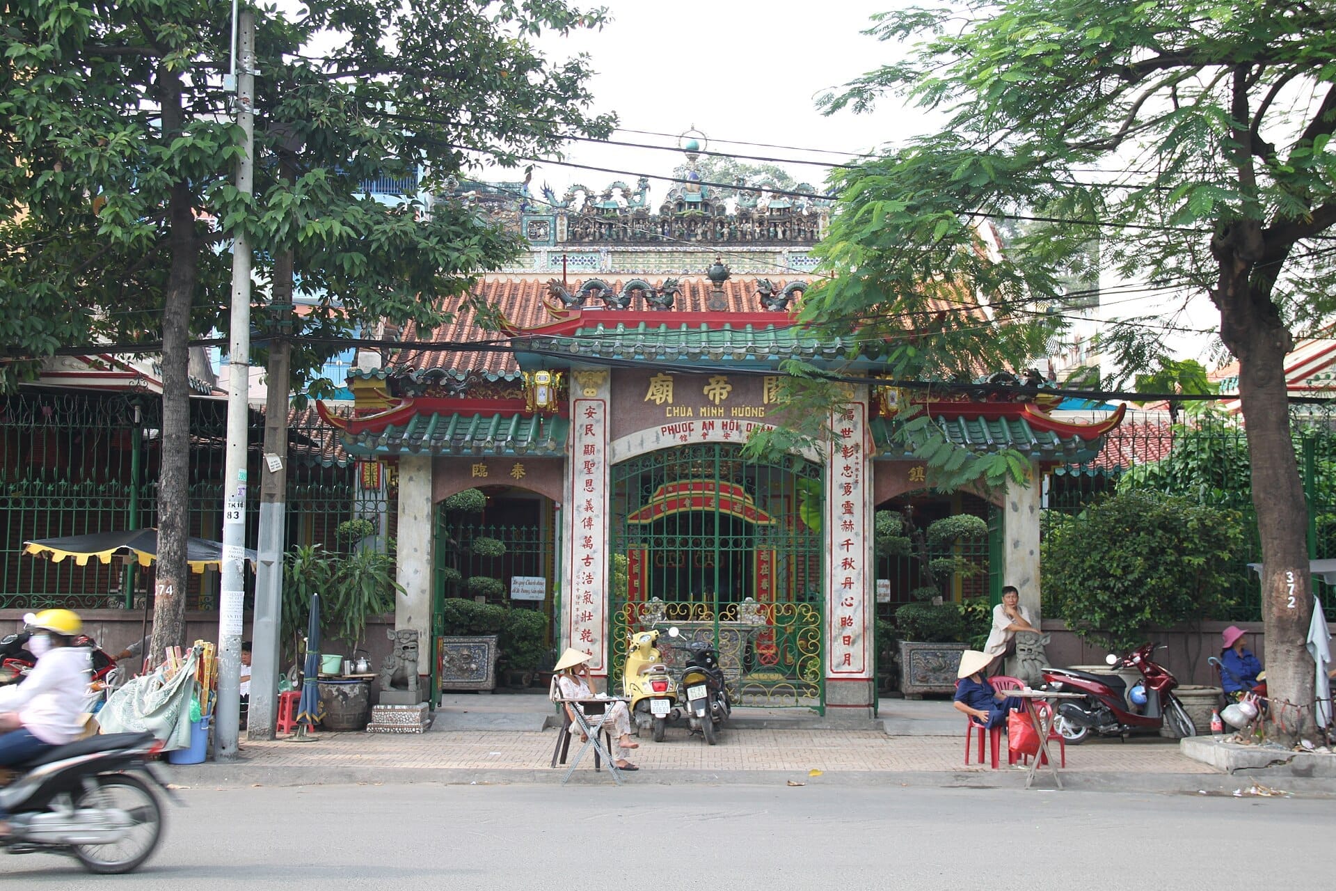 Vista frontal de la Pagoda Phước An Hội Quán con su ornamentado techo de tejas, puerta decorativa, plantas en macetas y personas sentadas afuera en una concurrida calle.