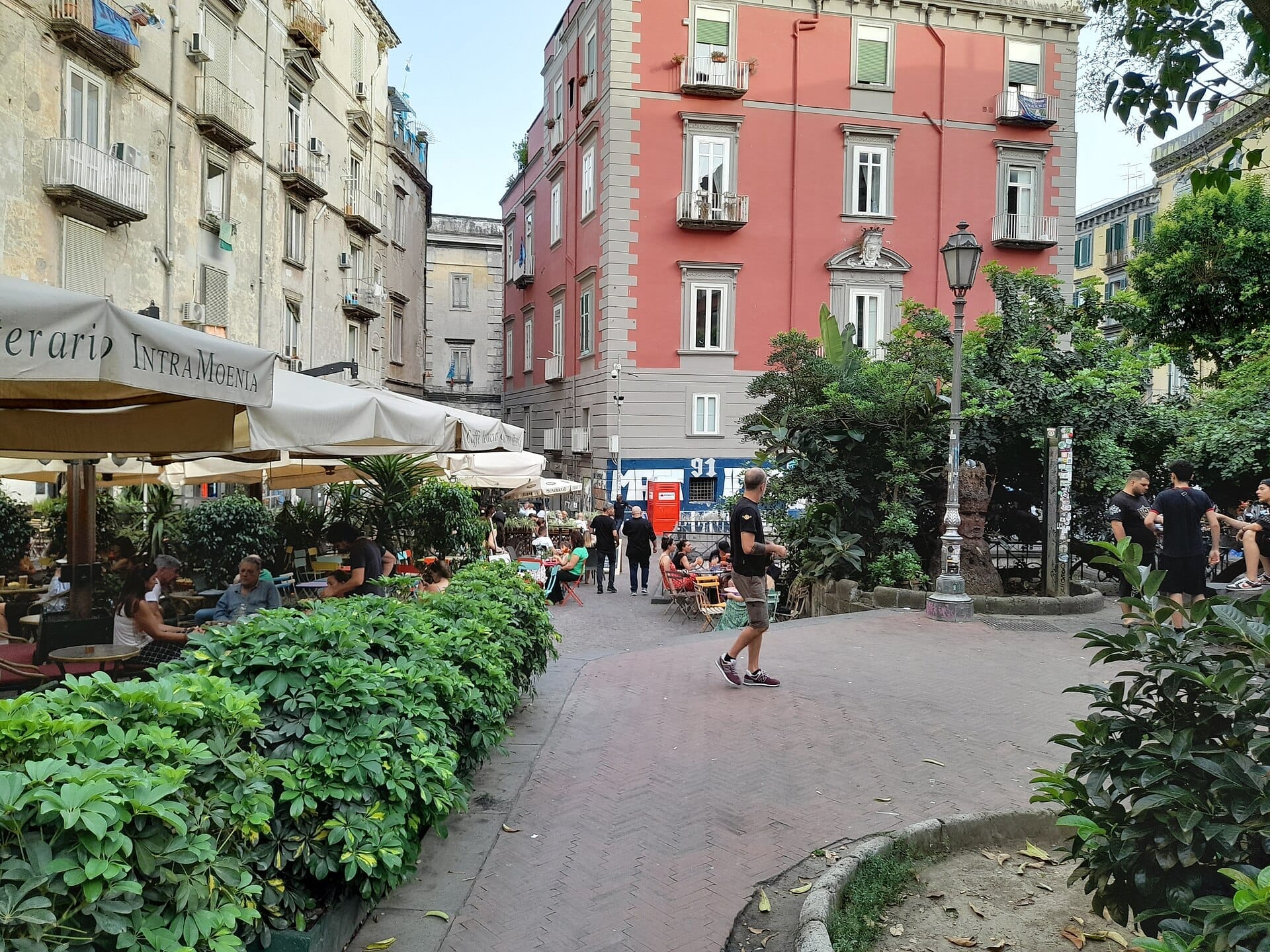 Vista di Piazza Bellini a Napoli con caffè all'aperto, rigogliosa vegetazione, edifici storici e persone che si godono la vivace piazza durante il giorno.