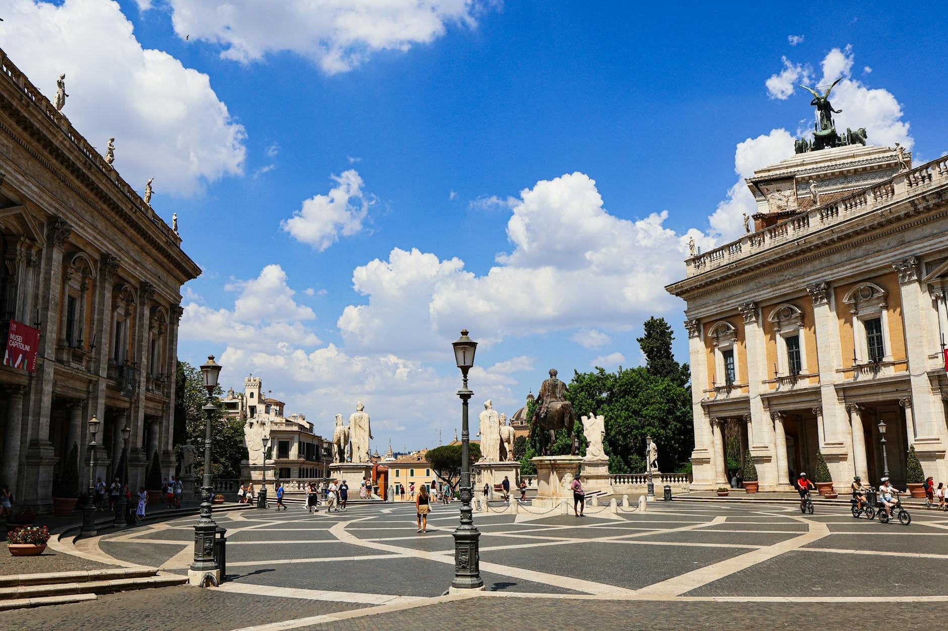 Wide view of Piazza del Campidoglio in Rome on a sunny day, showing Michelangelo’s geometric pavement, grand historic buildings, statues, and people enjoying the elegant square.
