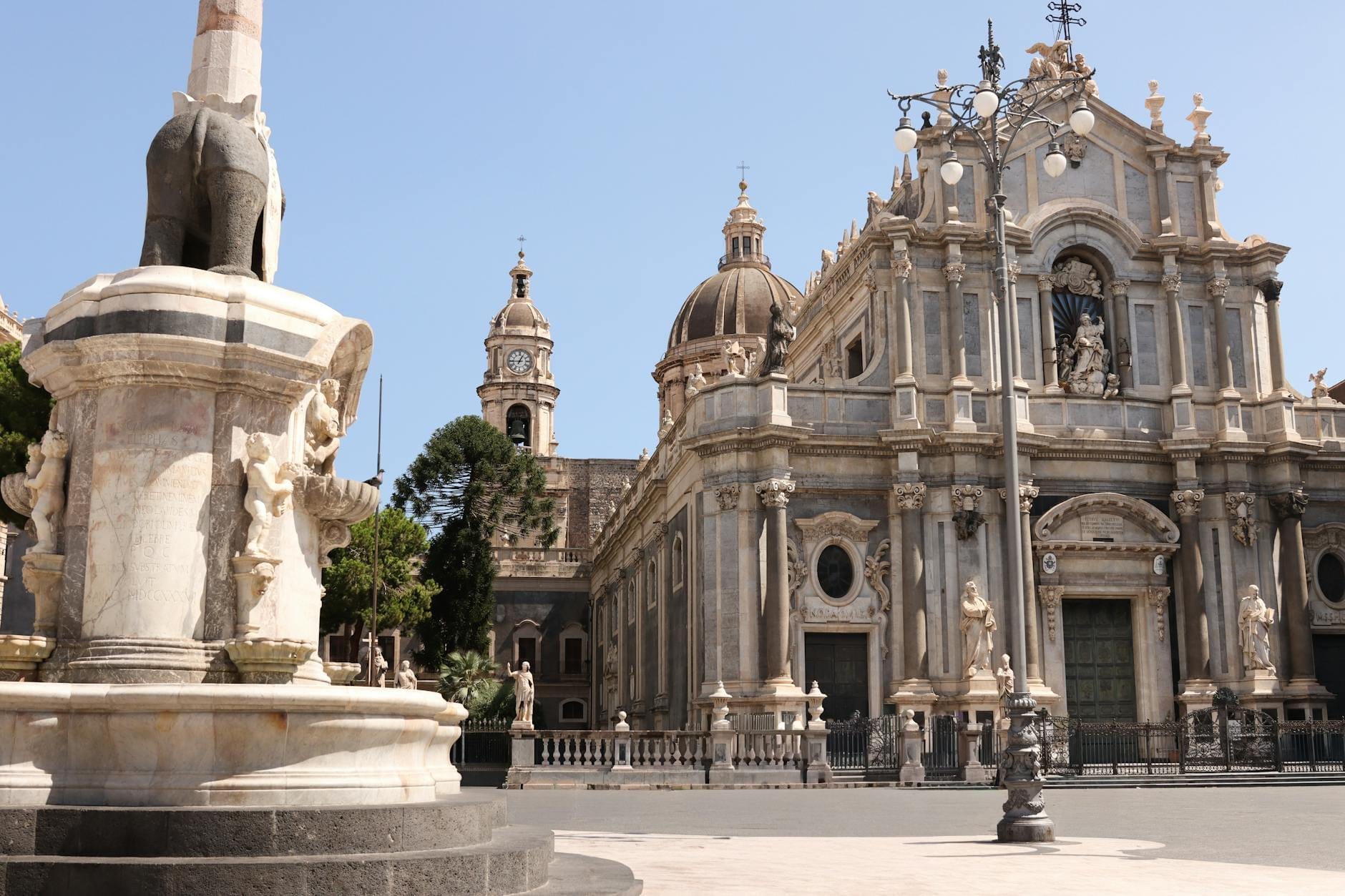 Blick auf die Piazza del Duomo in Catania mit dem ikonischen Elefantenbrunnen und der barocken Kathedrale von Catania vor klarem Himmel und mit prächtigen Architekturdetails.