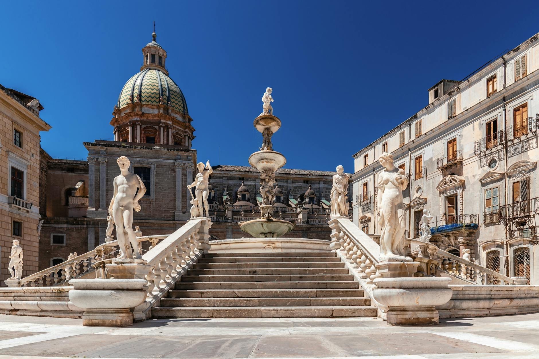 Weitwinkelansicht der großen Freitreppe der Piazza Pretoria und des Renaissance-Brunnens, umgeben von mythologischen Aktstatuen, mit historischen Gebäuden und strahlend blauem Himmel in Palermo, Sizilien.