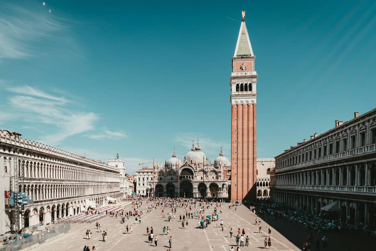 Weitwinkelbild der Piazza San Marco mit Campanile, Basilika di San Marco und den flankierenden Kolonnaden an einem klaren Tag mit Menschen auf dem Platz.