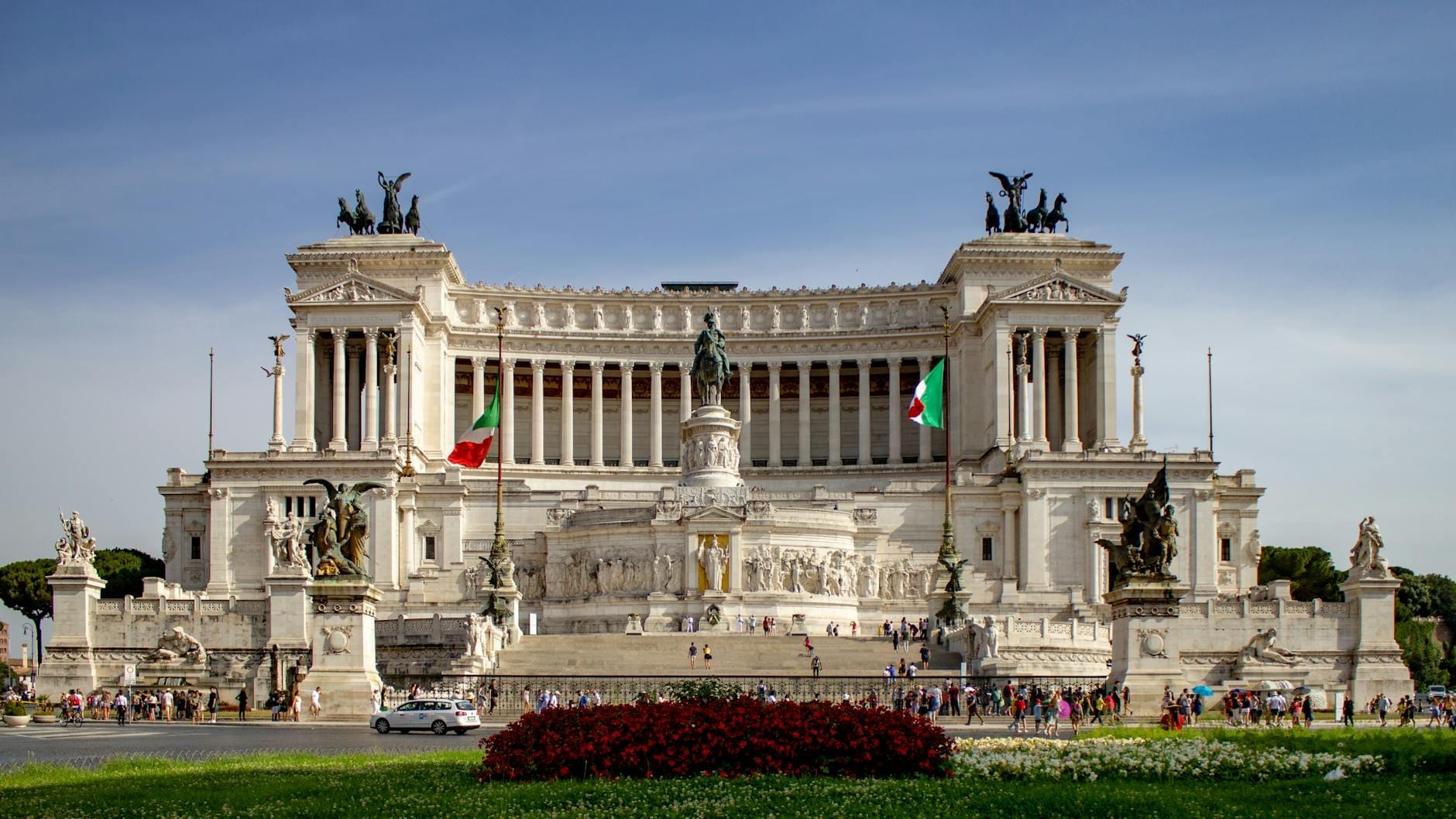 Wide, sunlit view of the Vittoriano monument at Piazza Venezia, Rome, with Italian flags, statues, and people gathered in front, under a clear sky.