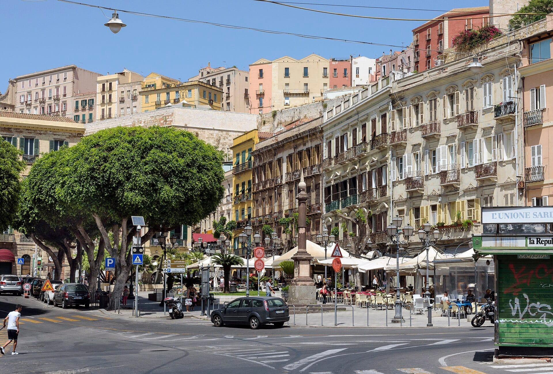 Piazza Yenne in Cagliari mit historischen Gebäuden, belaubten Bäumen, Straßenschildern, Cafés im Freien und einer Statue im Zentrum des Stadtplatzes.