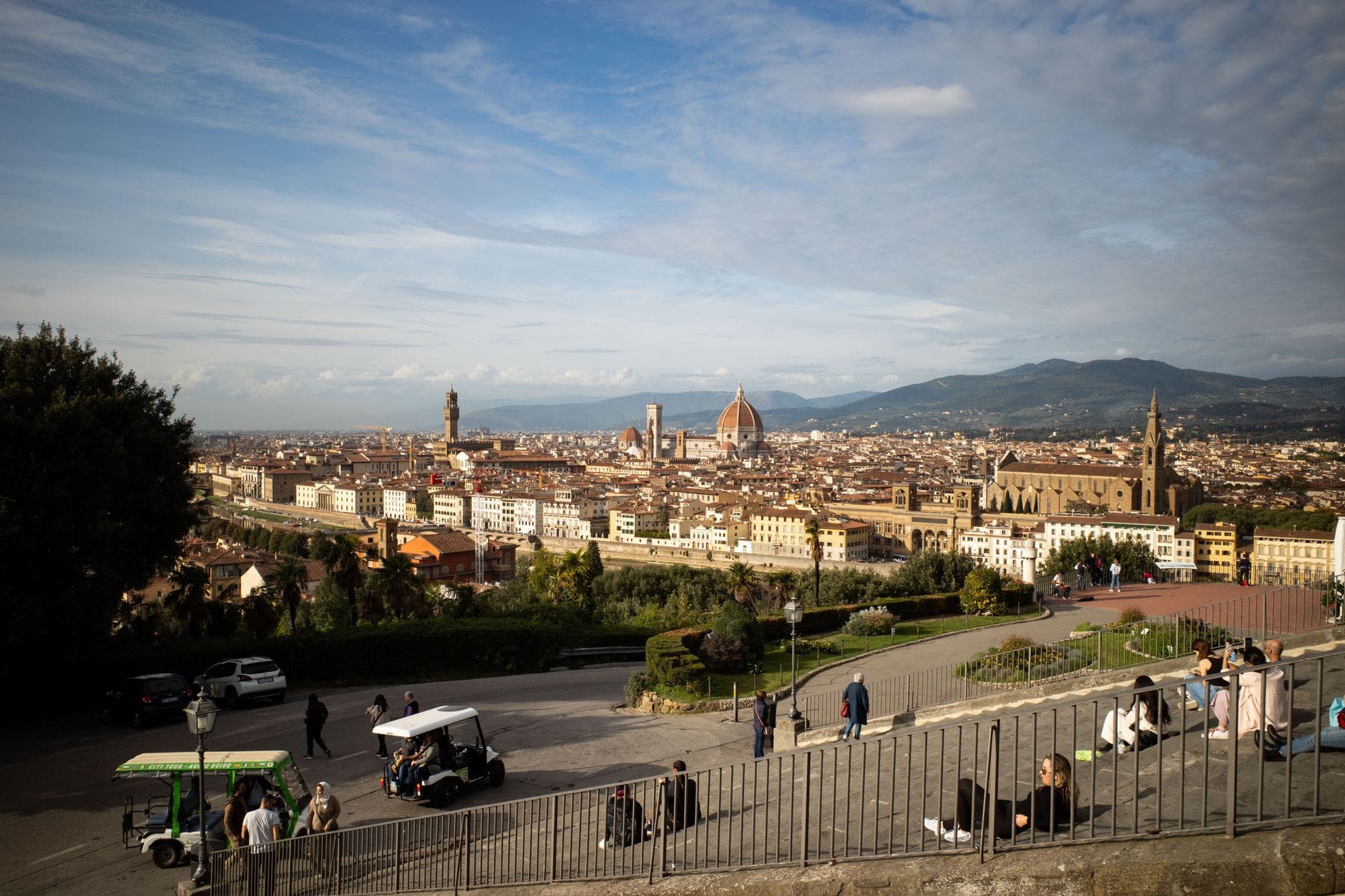 วิวกว้างจาก Piazzale Michelangelo เห็นเมืองฟลอเรนซ์ โดม Duomo หอคอย Palazzo Vecchio และแม่น้ำอาร์โน มีนักท่องเที่ยวและระเบียงอยู่ด้านหน้า