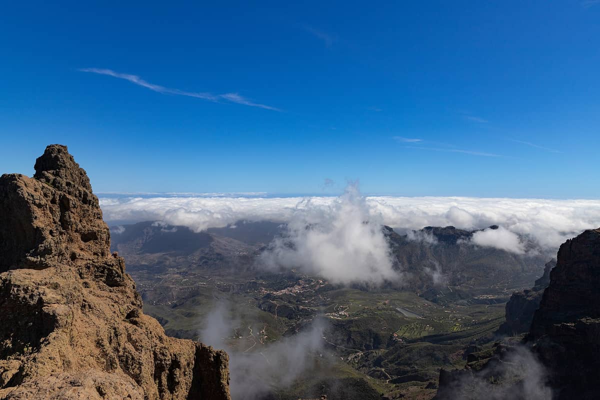 Панорамный вид с Pico de las Nieves: драматические скалистые обрывы, зелёные долины и море облаков под ярко-голубым небом.