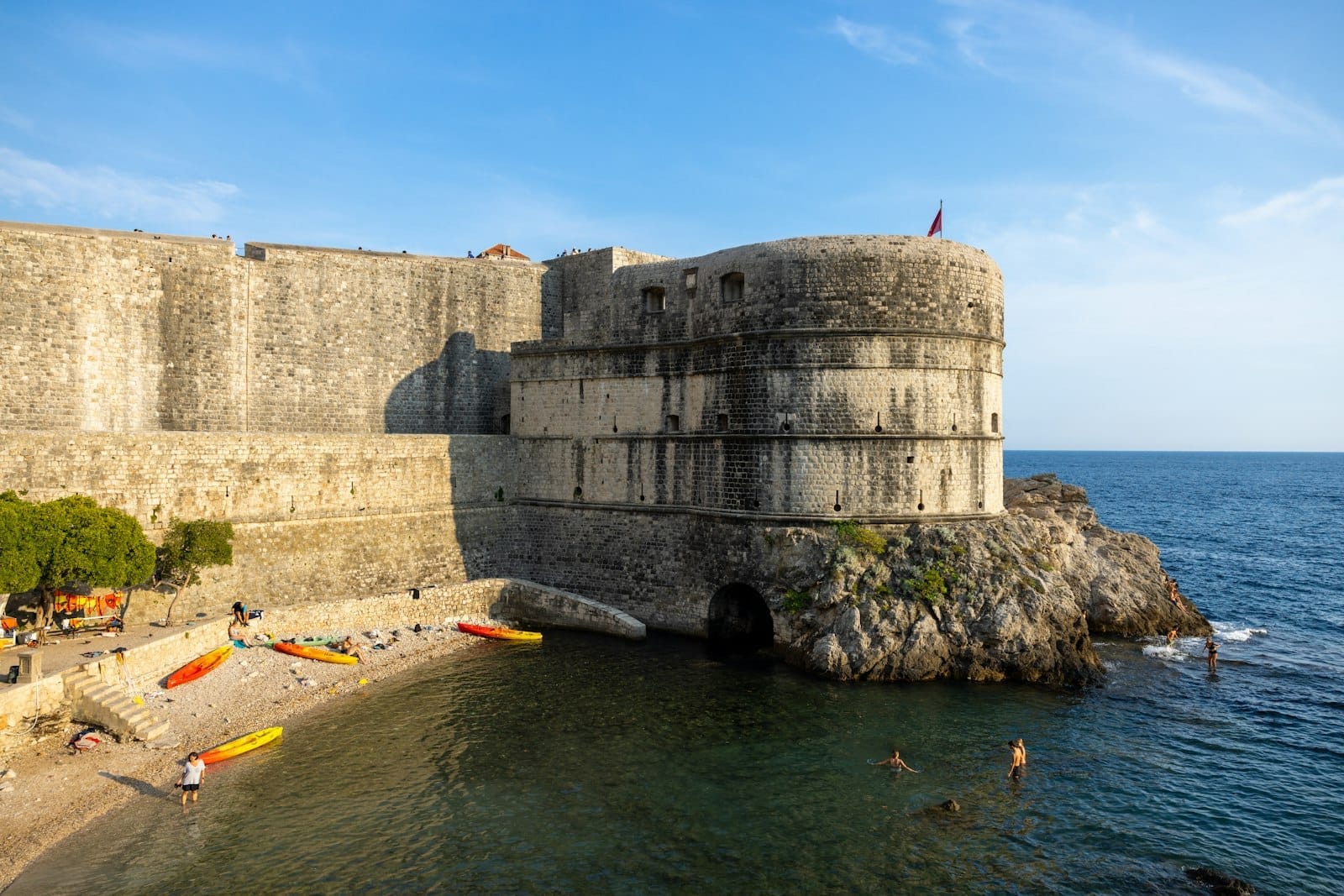 Vista de la muralla de la Puerta de Pile en Dubrovnik alzándose sobre el mar, con personas en la playa, kayaks y un cielo azul despejado.