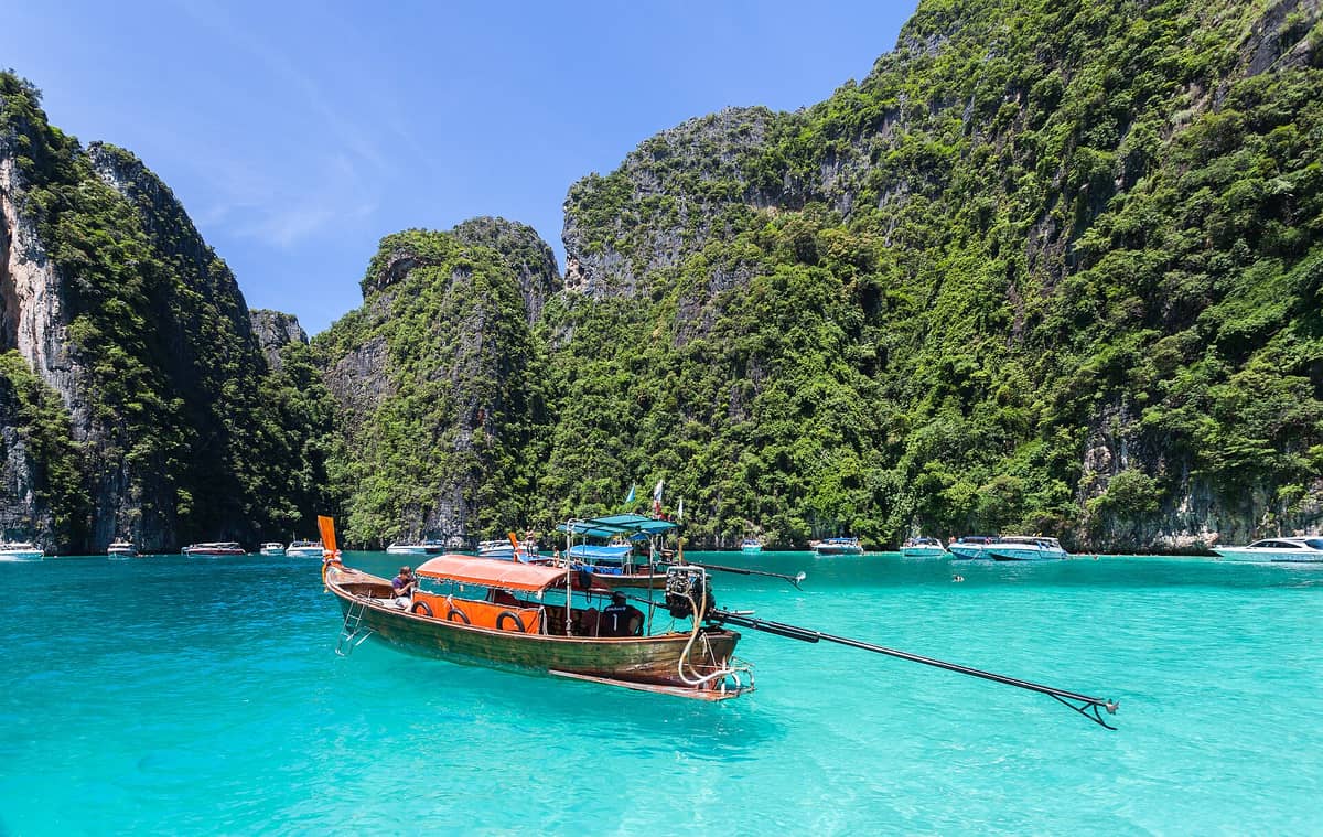 เรือหางยาวลอยอยู่กลางน้ำสีเขียวมรกตในลากูนปีเละ ล้อมรอบด้วยหน้าผาหินปูนสูงและต้นไม้เขียวขจี ใต้ท้องฟ้าสดใส