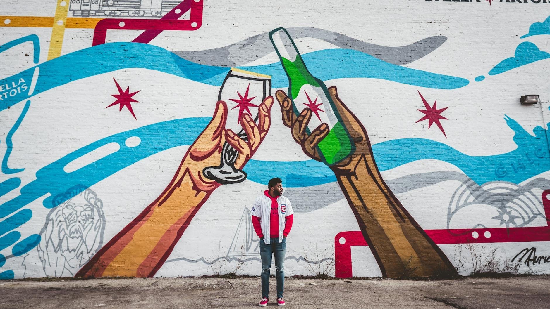 Un murale colorato su un muro raffigurante due grandi mani che fanno un brindisi con una bottiglia di birra e un bicchiere di vino, con una persona in piedi davanti. Stelle e onde in stile Chicago completano la vivace scena di street art.