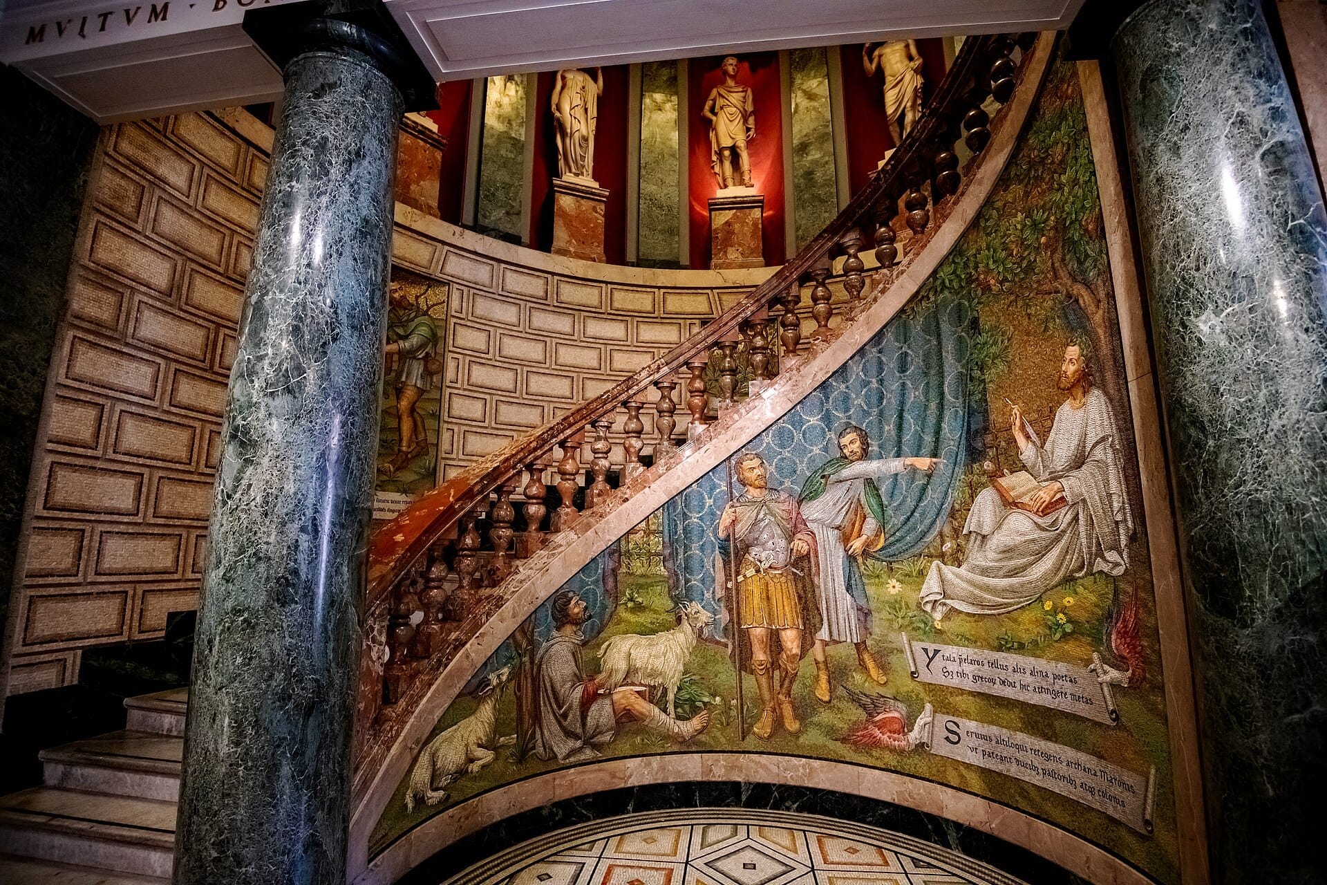 Scalinata in marmo con un vivido mosaico e statue classiche alla Pinacoteca Ambrosiana di Milano, con colonne ornate e arte parietale dettagliata.