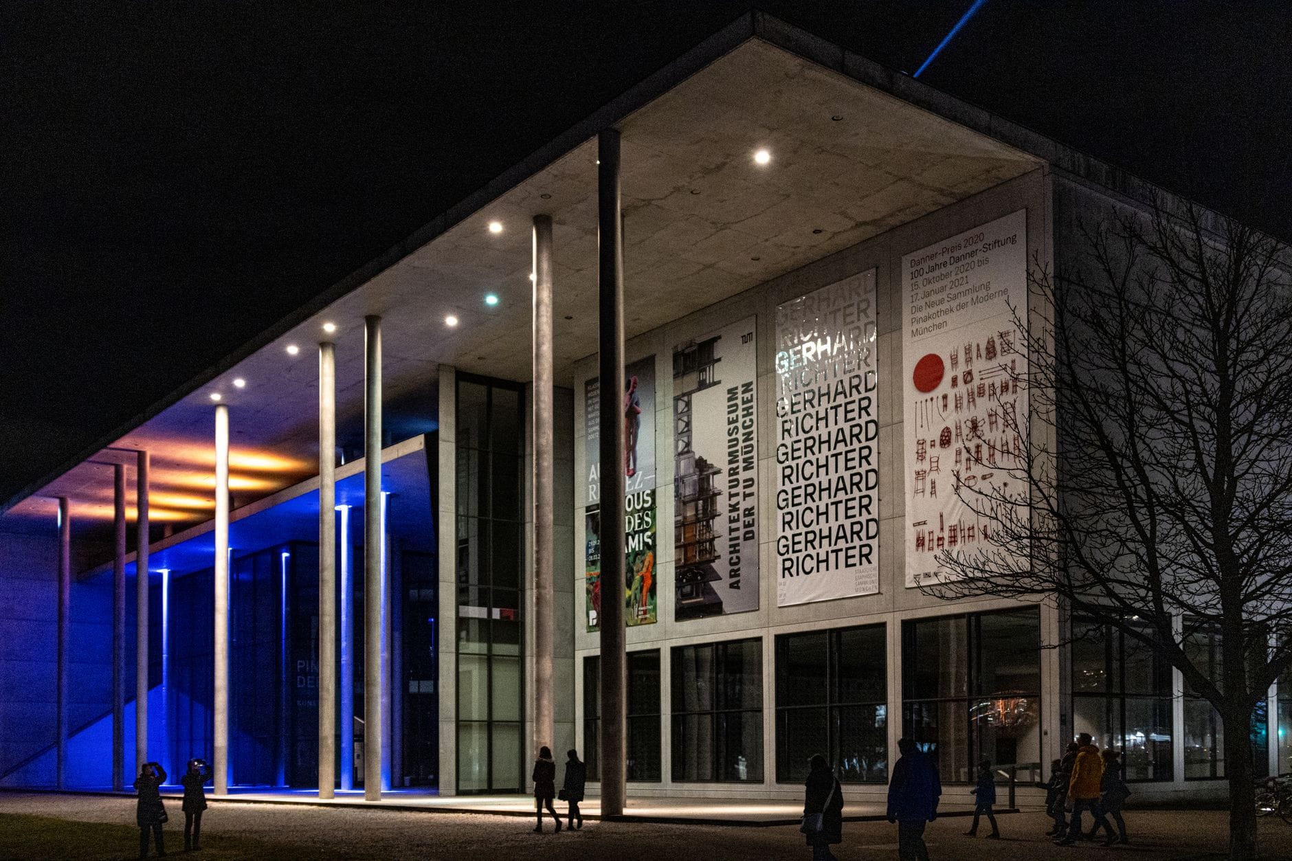 ภาพอาคารกระจกและคอนกรีตของ Pinakothek der Moderne กลางคืน ไฟน้ำเงินขาวสว่างตัดกับป้ายผลงานศิลปะโมเดิร์น