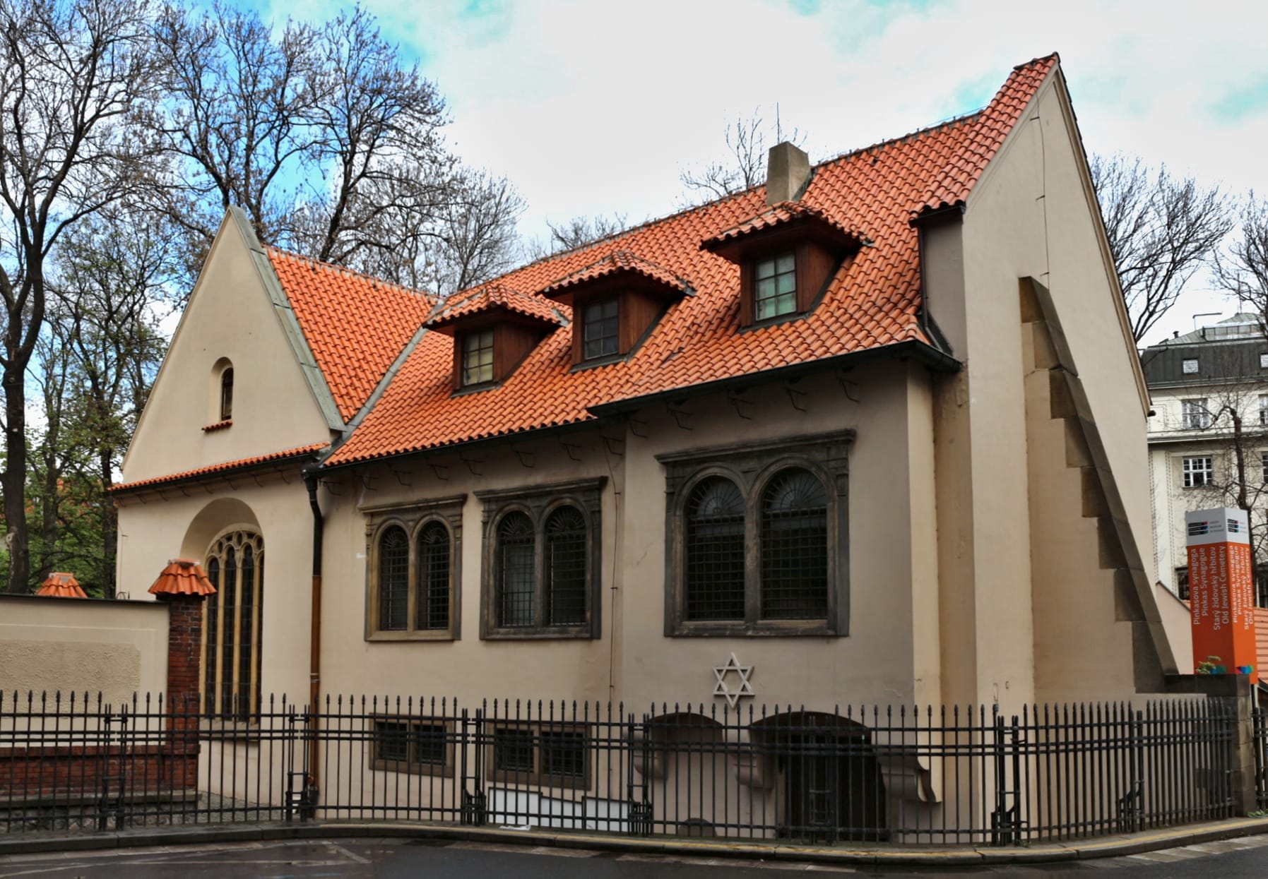 Widok zewnętrzny Synagogi Pinkasa w Pradze, z kremowymi ścianami, czerwonym dachówkowym dachem, łukowymi oknami i Gwiazdą Dawida nad wejściem.
