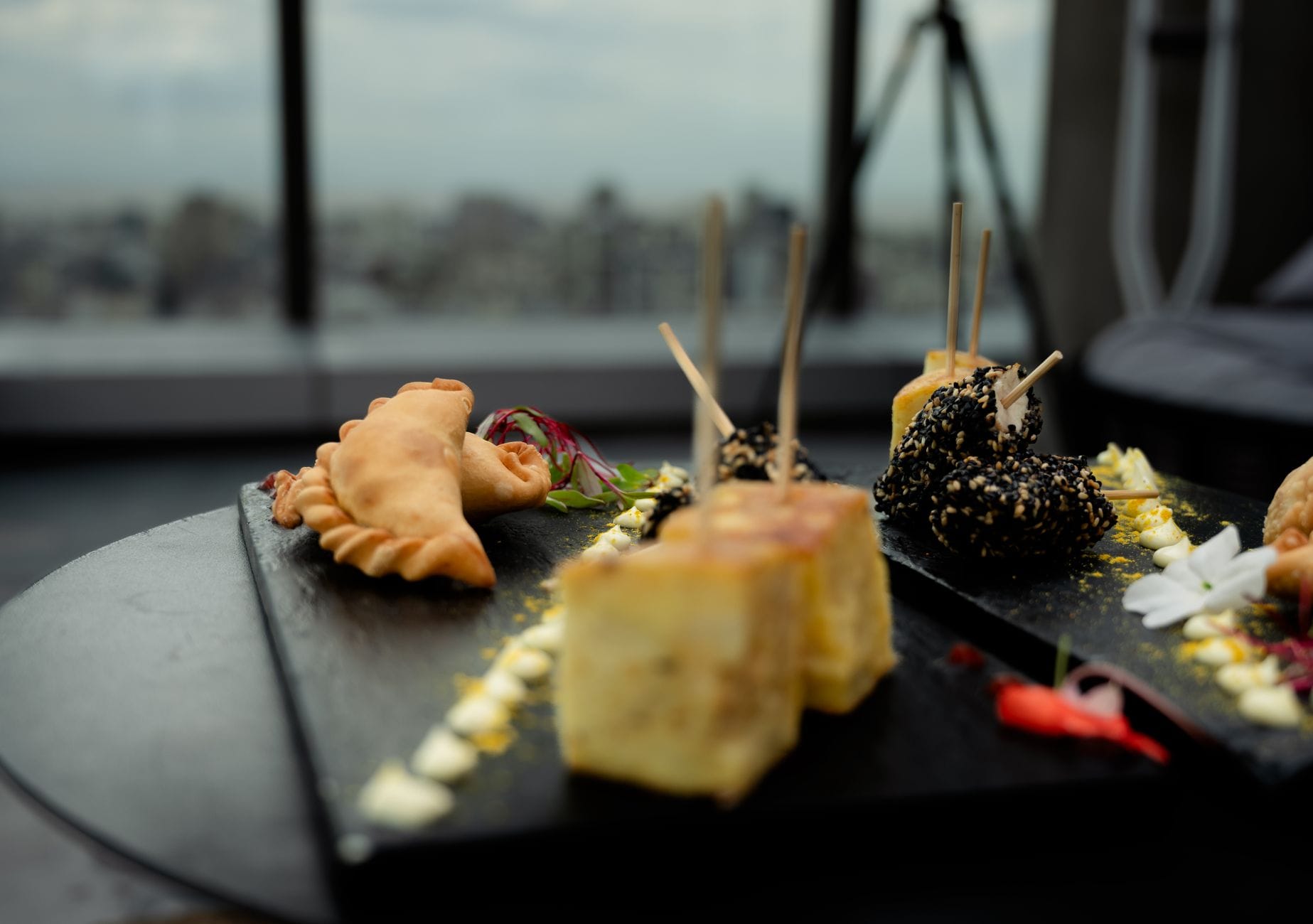 Close-up of assorted pintxos on black slate trays, featuring tortilla, empanada, sesame skewers, and garnishes in a bar with a city view background.