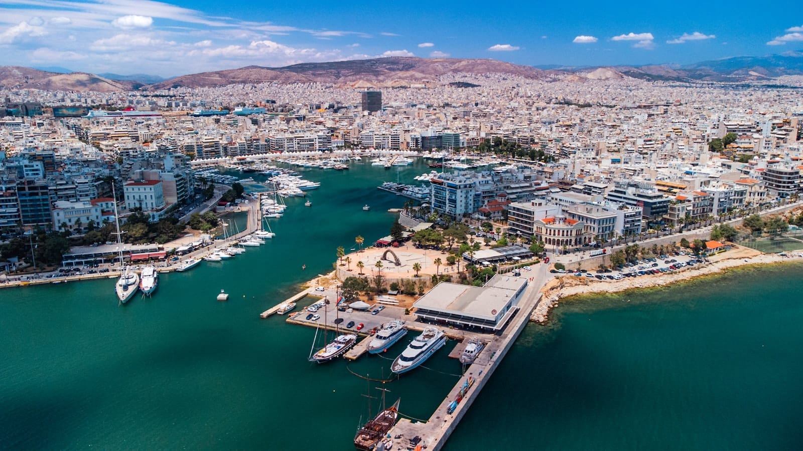 มุมมองทางอากาศของท่าเรือ Piraeus พร้อมน้ำสีเทอร์คอยซ์ เรือยอชต์จอดเทียบท่า อาคารสมัยใหม่ และเนินเขาเป็นฉากหลังใต้ท้องฟ้าสีฟ้า