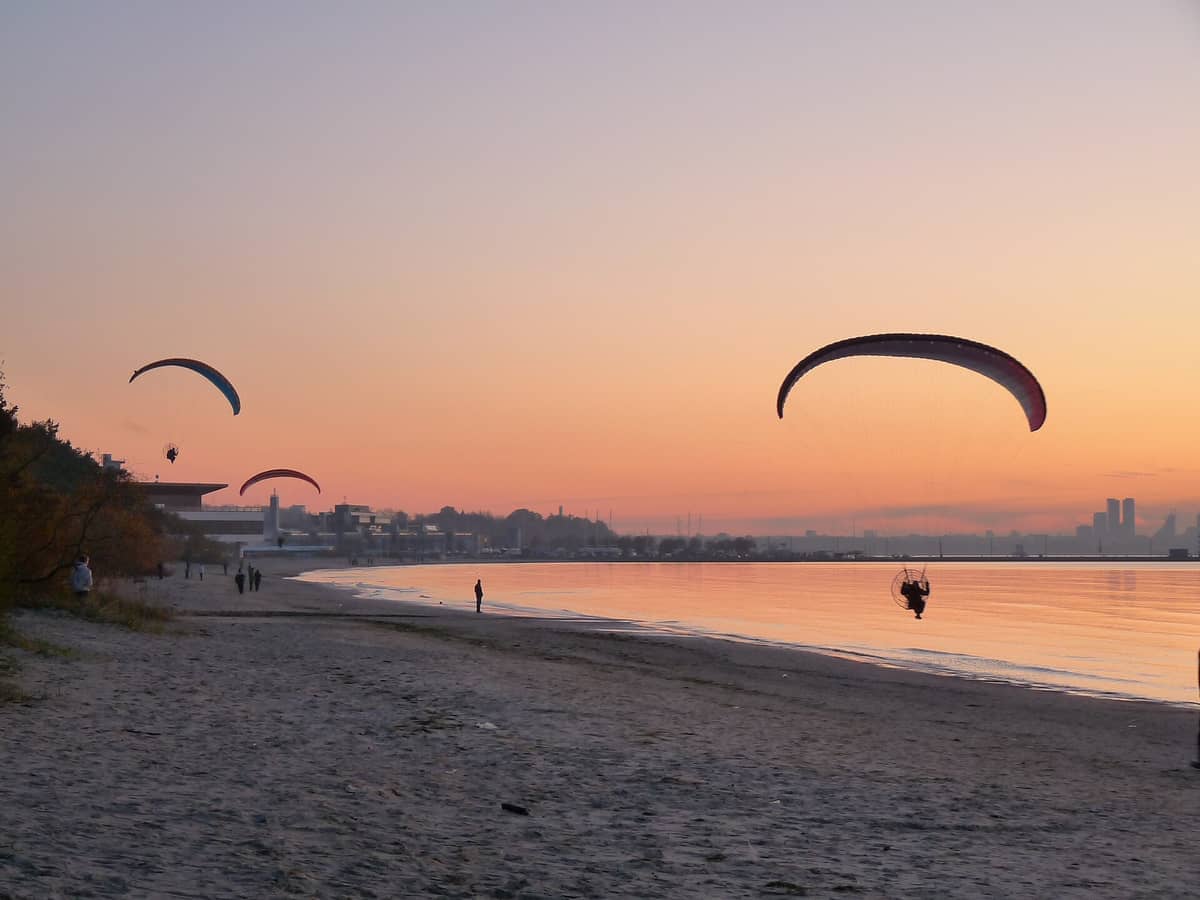 Gleitschirmflieger schweben bei Sonnenuntergang über dem Pirita Beach, mit ruhigem Wasser, Sandstrand und fernen Gebäuden unter einem Pastellhimmel.