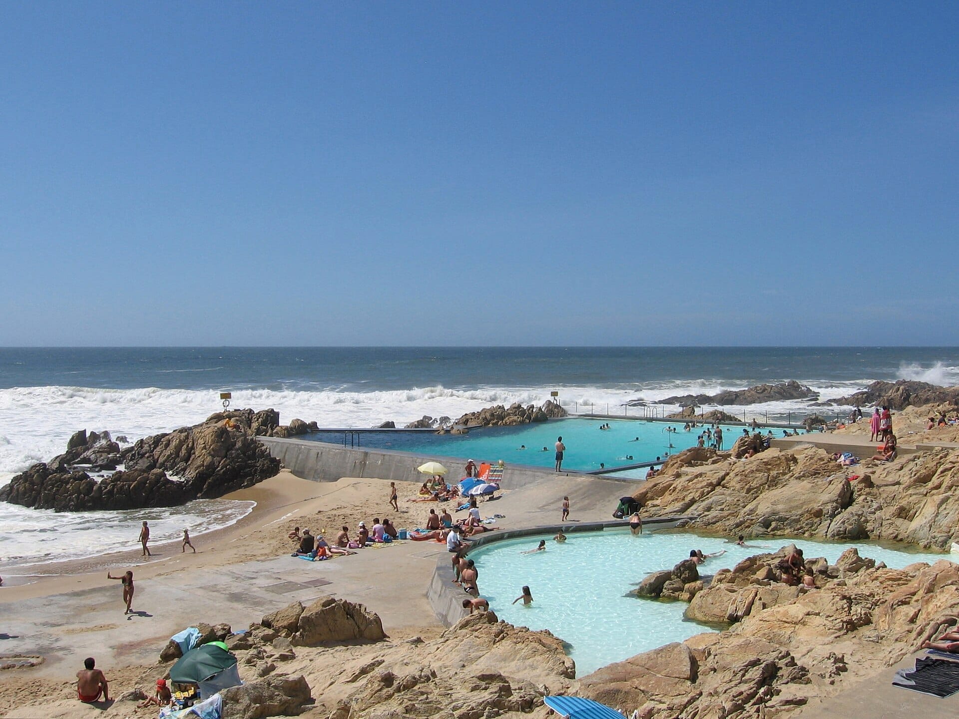 Le piscine di Piscina das Marés si fondono perfettamente con la costa atlantica rocciosa, con bagnanti, nuotatori e pietra naturale sotto un cielo azzurro limpido.