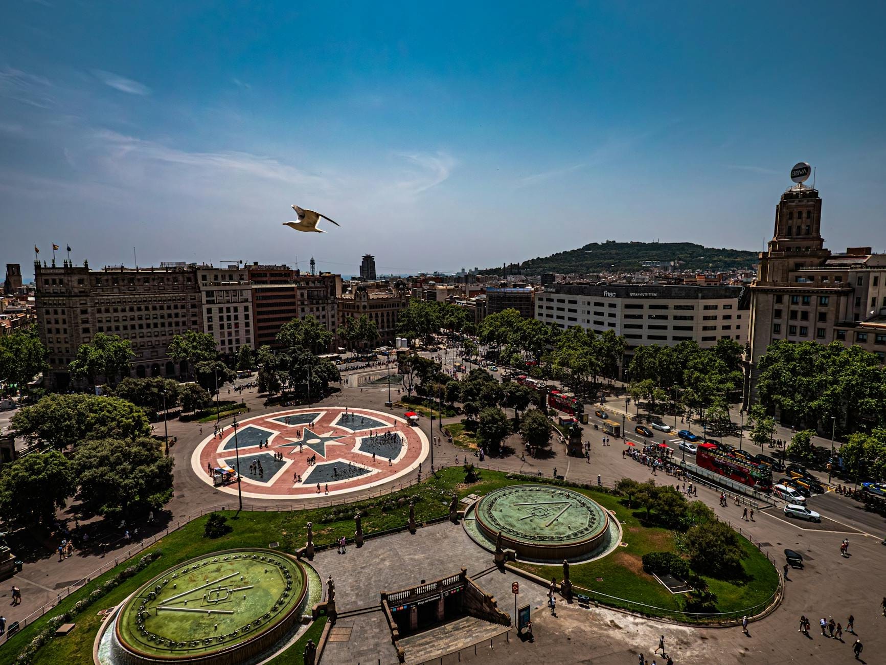 Aerial view of Plaça de Catalunya with its distinctive circular plaza, surrounding gardens, nearby buildings, and people enjoying the vibrant public square in Barcelona.