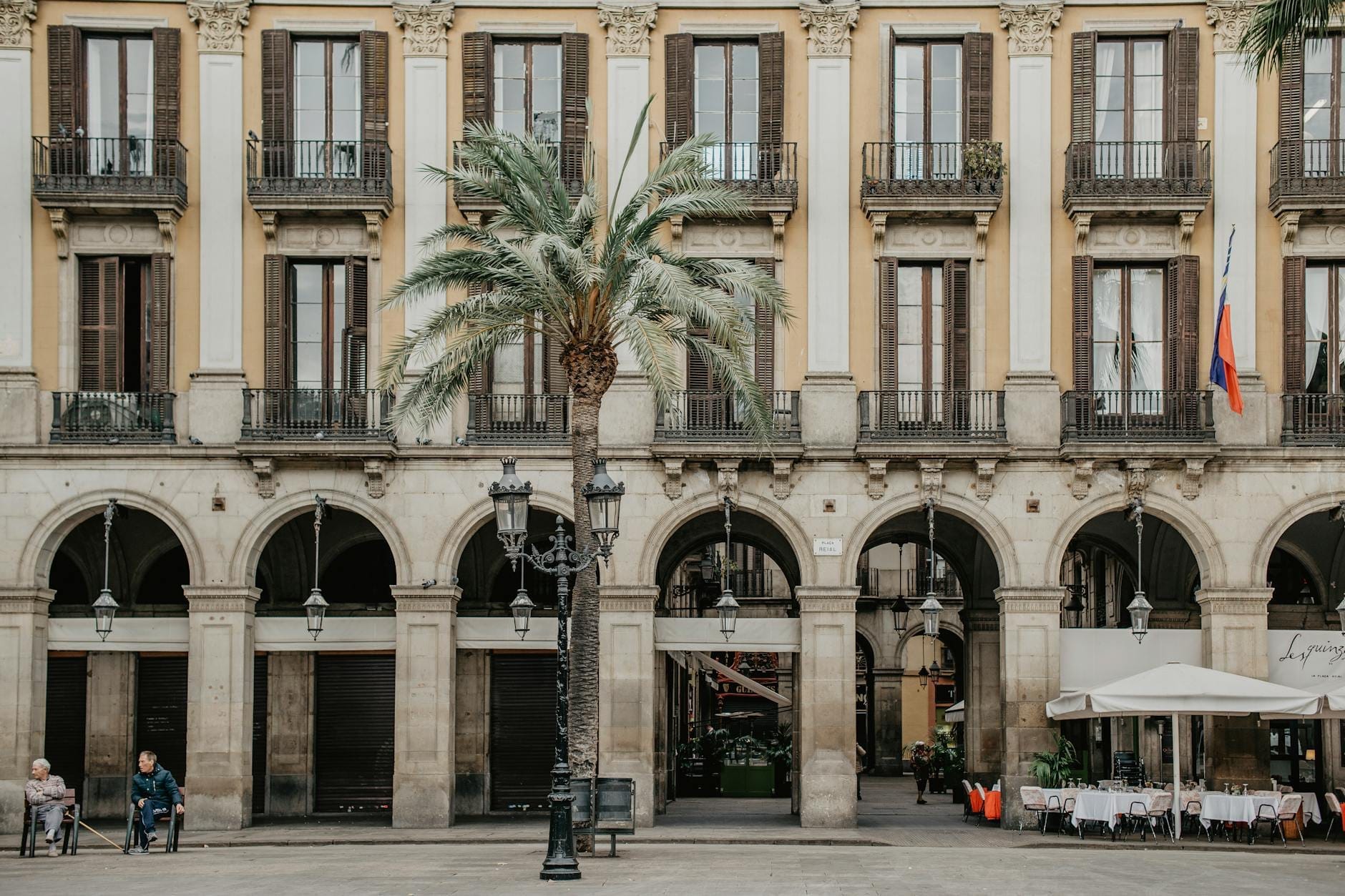 Arcaded yellow neoclassical buildings with balconies and palm trees in Plaça Reial, Barcelona, outdoor restaurant seating and people sitting in the square.