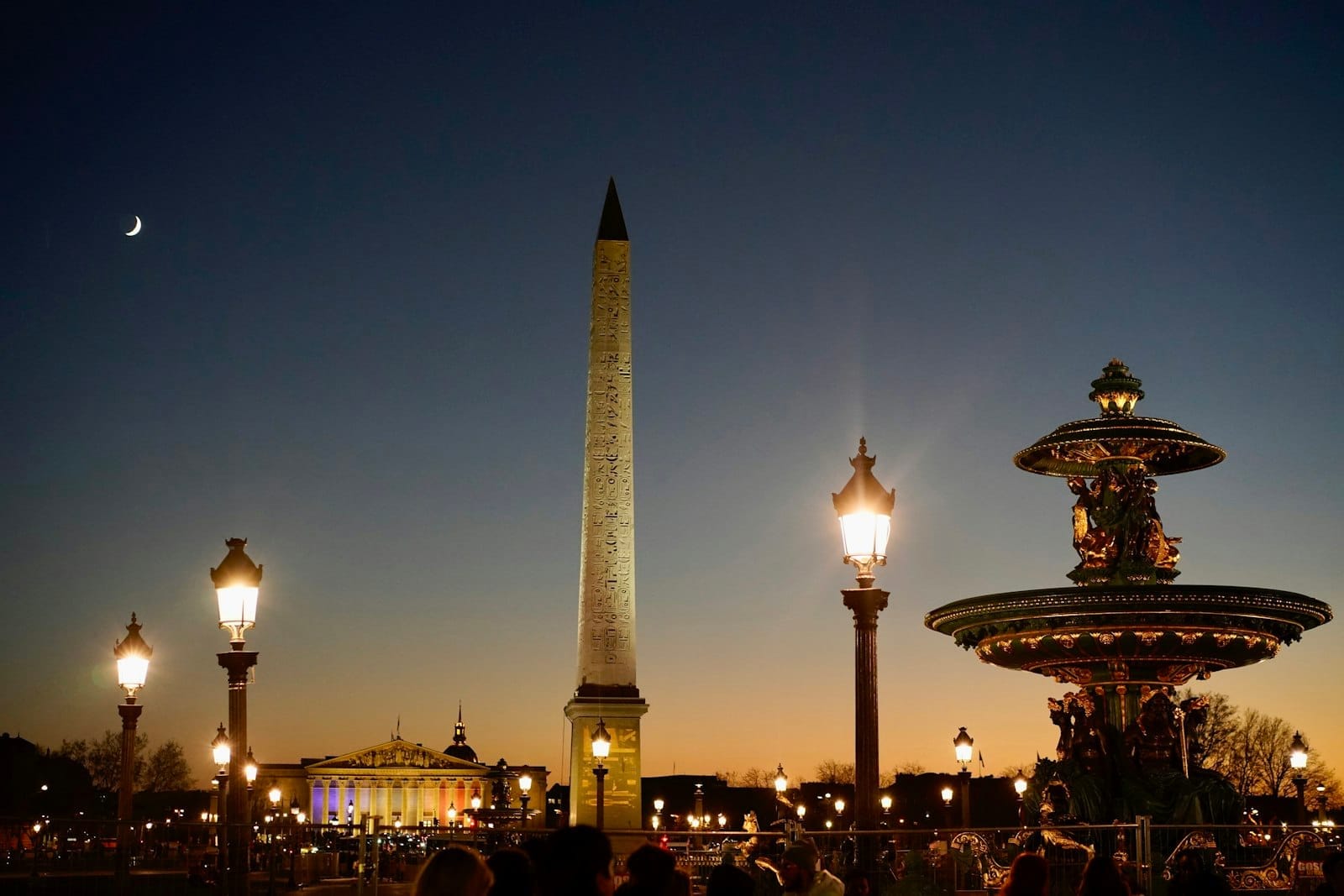 Place de la Concorde al crepuscolo con lampioni illuminati, l'obelisco egizio, una fontana monumentale e persone che animano la piazza sotto un cielo al tramonto.