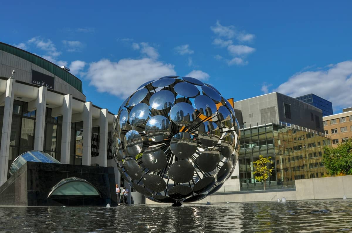 Eine große, reflektierende kugelförmige Skulptur steht vor dem Place des Arts in Montreal, umgeben von moderner Architektur unter einem strahlend blauen Himmel.
