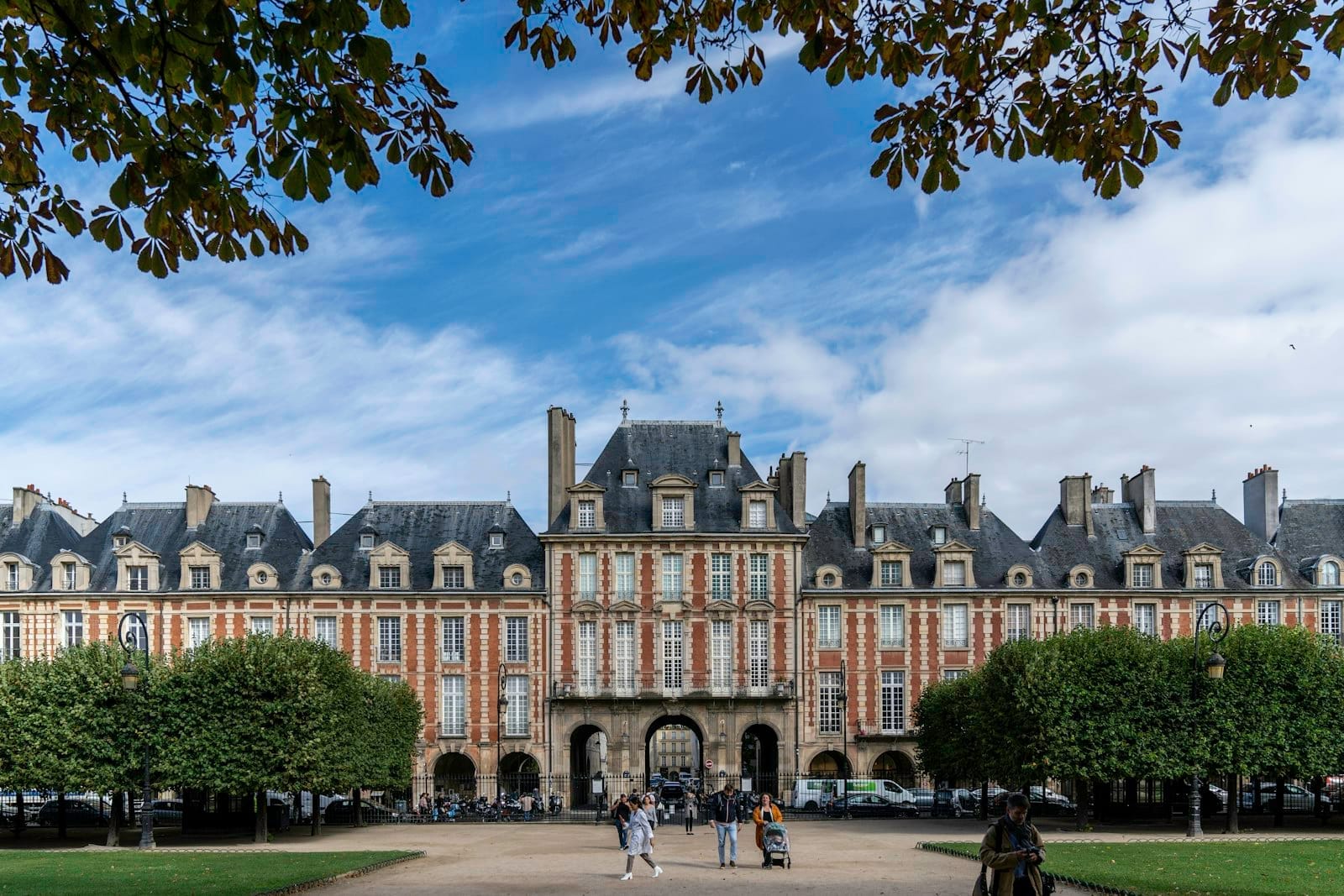 Veduta ampia della Place des Vosges con i padiglioni in mattoni rossi, il giardino formale, gli alberi e le persone che passeggiano sotto un cielo azzurro con qualche nuvola.